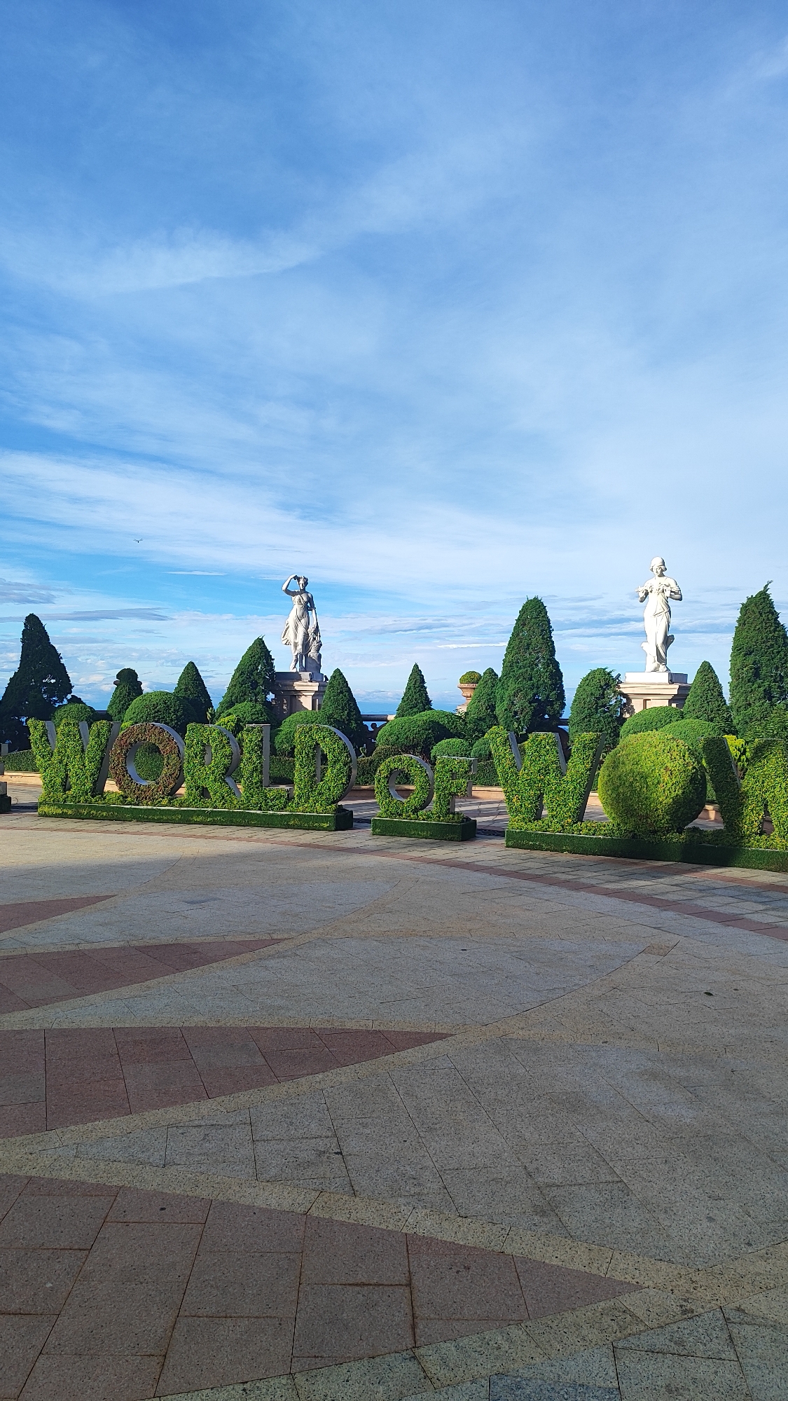 The image shows a park with a large topiary sculpture that spells out "WORLD OF JOY." The letters are made of green bushes and are arranged in rows. Behind the sculpture are several stone statues of women in classical attire, as well as well-manicured cypress trees and other ornamental plants. The ground in front of the sculpture is paved with patterned tiles in shades of beige and terracotta. The sky above is blue with scattered white clouds. This scene is located at the Asia Park in Da Nang, Vietnam.