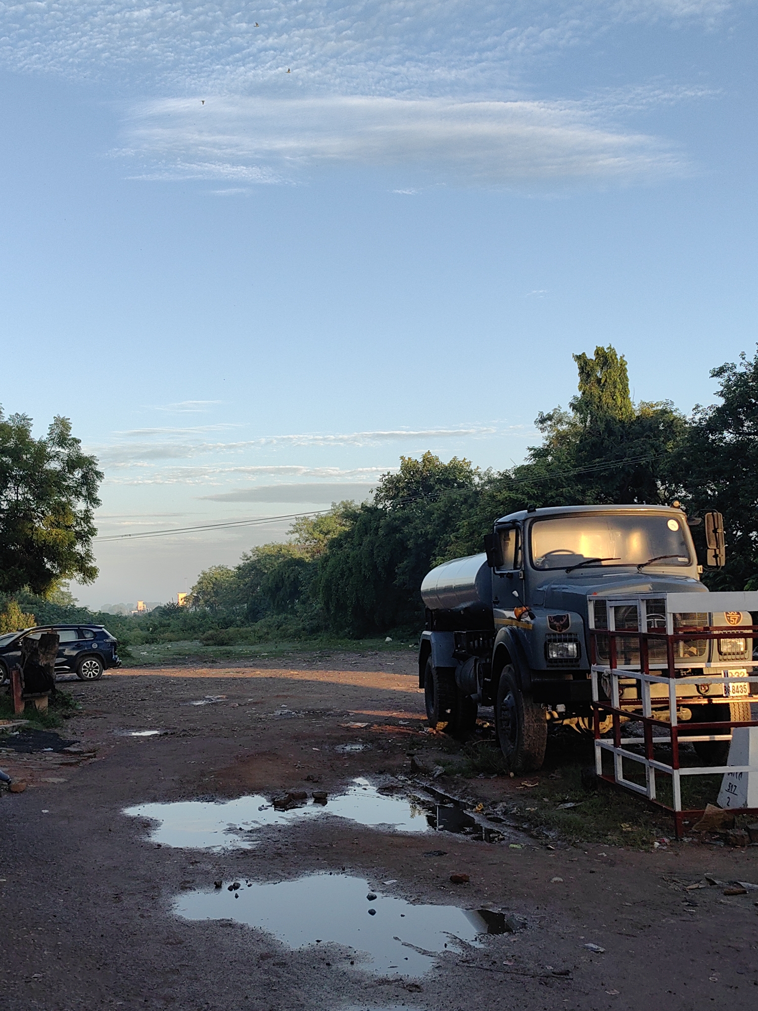 In Belahiya, Nepal, a gray water tanker truck is parked on a muddy, unpaved road. Puddles of water are present on the ground. To the left of the truck, a dark-colored SUV is parked. The scene is illuminated by the soft light of what appears to be sunrise or sunset, casting a warm glow on the truck's windshield and the surrounding environment. Tall green trees line the background. The sky is a clear blue with scattered, wispy clouds and a few birds are visible in flight.