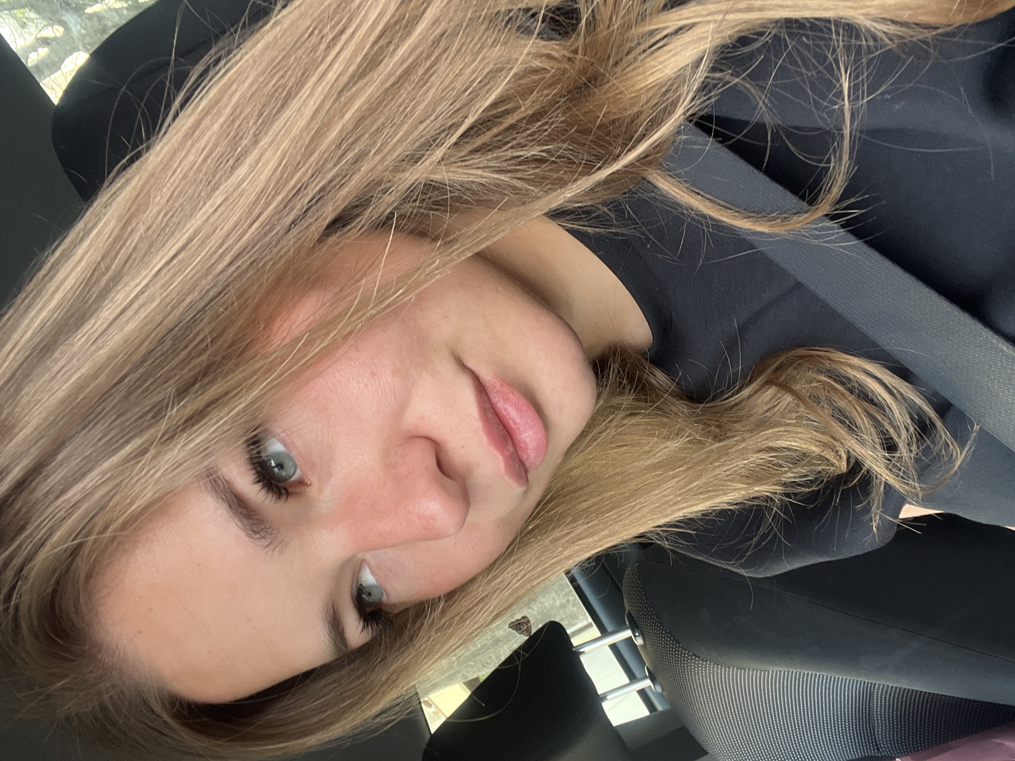 A young woman with long blonde hair is seated in a car, wearing a black shirt and a seatbelt. The interior of the car is visible, including the seats and a window. The background is blurred, but hints at an outdoor environment. It is not possible to definitively confirm the location as Budva, Montenegro, based solely on this image.
