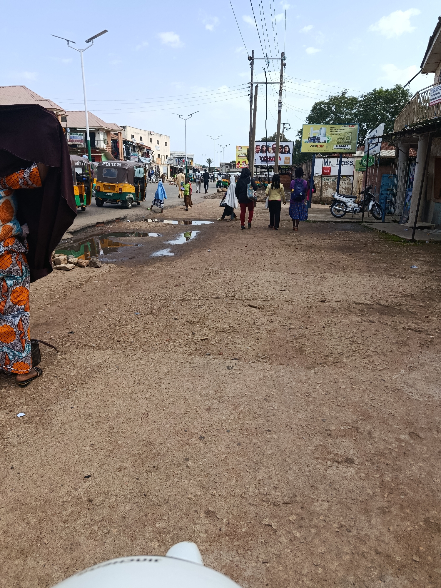 The image depicts a street scene in Jos, Nigeria. In the foreground, a person is partially visible on the left, wearing a colorful patterned skirt and a dark head covering. The street itself is unpaved and dusty, with several puddles of water scattered across it. To the left, a yellow and green auto-rickshaw (locally known as a 'Keke Napep') is parked or moving. Several other people are walking along the street, including a group of women in the middle ground.
In the background, there are buildings lining the street, with shops and businesses visible. Billboards are erected along the road, one of which advertises "Mexe for Household Appliances" and another displays faces with the text "YEHUM". A white motorcycle is parked on the right side of the street near a shop. Overhead, power lines crisscross the sky, supported by utility poles. The sky is partly cloudy. The overall impression is of a bustling, everyday street scene in an urban or semi-urban area of Jos.
