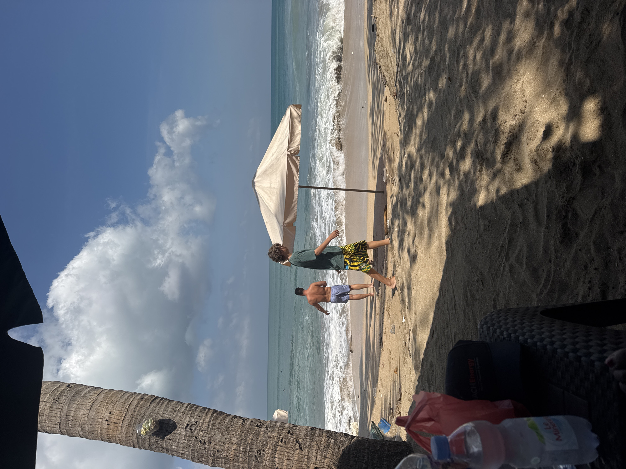On a sunny day in Kuta, Indonesia, the turquoise ocean laps at a sandy beach. Two young men are enjoying the water. One, wearing blue swim trunks, stands facing the ocean. The other, in colorful swim trunks, walks away from the water towards a beige umbrella that offers shade on the sand. A palm tree stands tall to the left of the frame, its trunk textured and its leaves casting shadows on the ground. In the foreground, a plastic chair holds a bottle of water and a red plastic bag. The sky is a bright blue, dotted with fluffy white clouds.