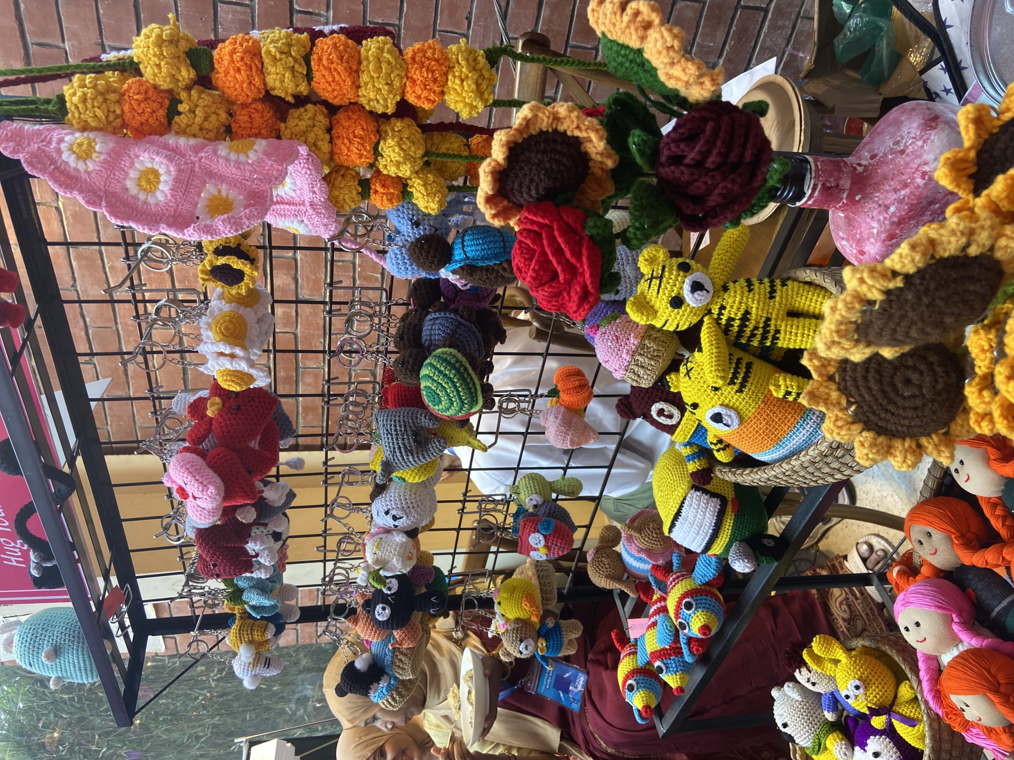 The image shows a collection of handcrafted items, likely for sale at a market or fair. The items, mostly crocheted, include flower garlands (marigolds and sunflowers), small animal keychains (tigers, bears), and dolls. They are displayed on a metal grid structure and in baskets. At least one person with a head covering is visible beneath the display. The context, based on the location, is likely an artisan market in Dhaka, Bangladesh. The scene suggests a focus on local crafts and handmade goods.