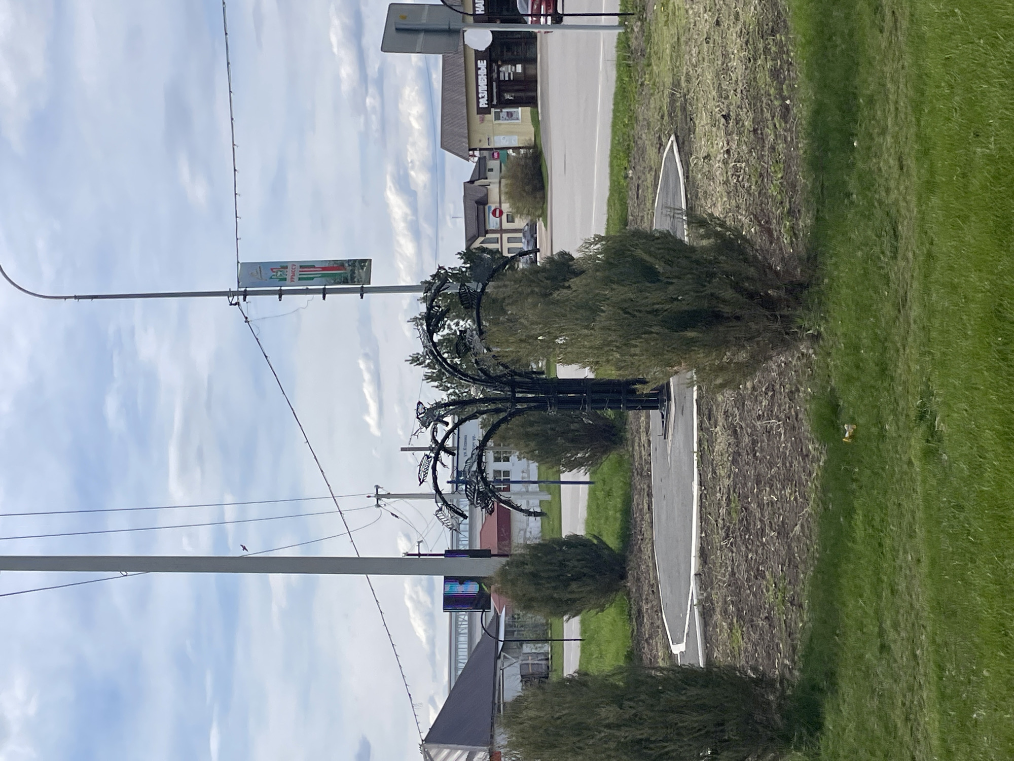 The image captures a street scene in Urussu, Russia. A sidewalk runs alongside a road, with patches of grass on either side. Small, rounded bushes line the sidewalk. A decorative, wrought-iron structure, resembling a stylized tree, stands along the sidewalk. Overhead, power lines stretch across the sky. A building with a sign in Russian is visible in the background. The sky is partly cloudy. A sign with green, white, and red colors is attached to the pole on the left.