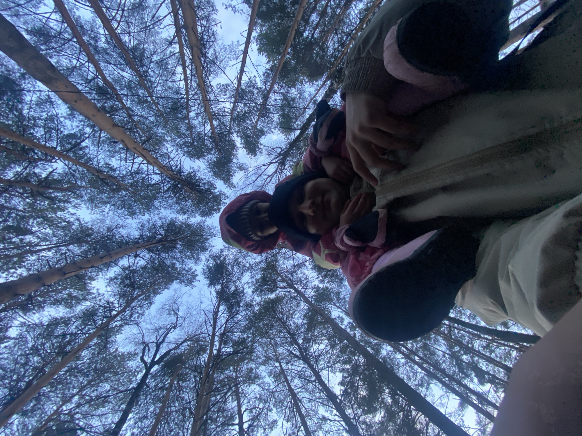 Two people are looking into the camera from a low angle against a background of tall trees in a forest. The sky is visible through the tree branches. The location is likely in the city of Chaikovsky, Russia. One person is wearing a red hooded jacket, and the other is wearing a light-colored jacket. One person has their hand near the zipper of a jacket.