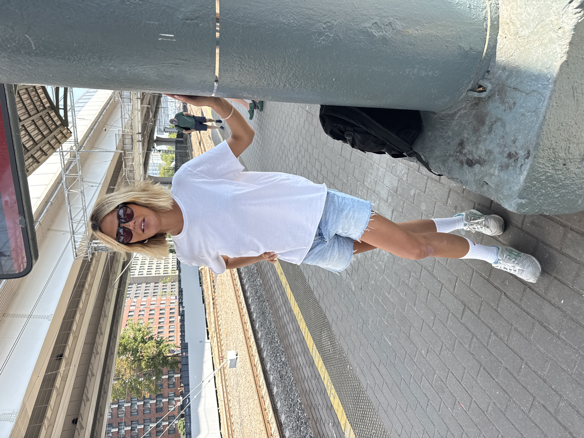 A woman with blonde hair and sunglasses stands on a train platform in Khimki, Russia. She is wearing a white t-shirt, denim shorts, white socks, and grey sneakers. Her left hand is on her hip, and her right arm is resting on a large grey pillar. A black backpack is on the ground next to the pillar. The platform is made of grey paving stones, with a yellow line marking the edge. Train tracks are visible behind the woman, and buildings and trees are in the background. The sky is blue and clear.