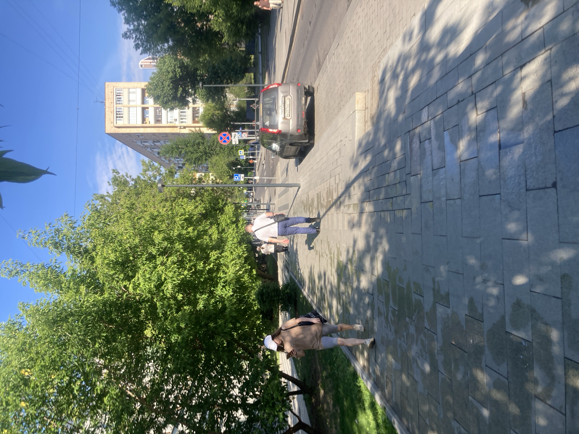 The image depicts a sunlit street scene in Moscow, Russia, characterized by a wide paved sidewalk lined with mature trees. A woman wearing a white cap, beige t-shirt, and grey cycling shorts walks on the sidewalk, holding a smartphone. Behind her, a man in a white shirt and blue pants walks away from the camera, carrying a dark shoulder bag. In the background, a grey SUV is parked on the street, and beyond that, a multi-story apartment building stands under a clear blue sky with some electrical wires visible. The sidewalk is wet in patches, suggesting recent rain or street cleaning, and dappled with shadows cast by the trees. Street signs, including a parking sign and a traffic sign, are visible near the road.