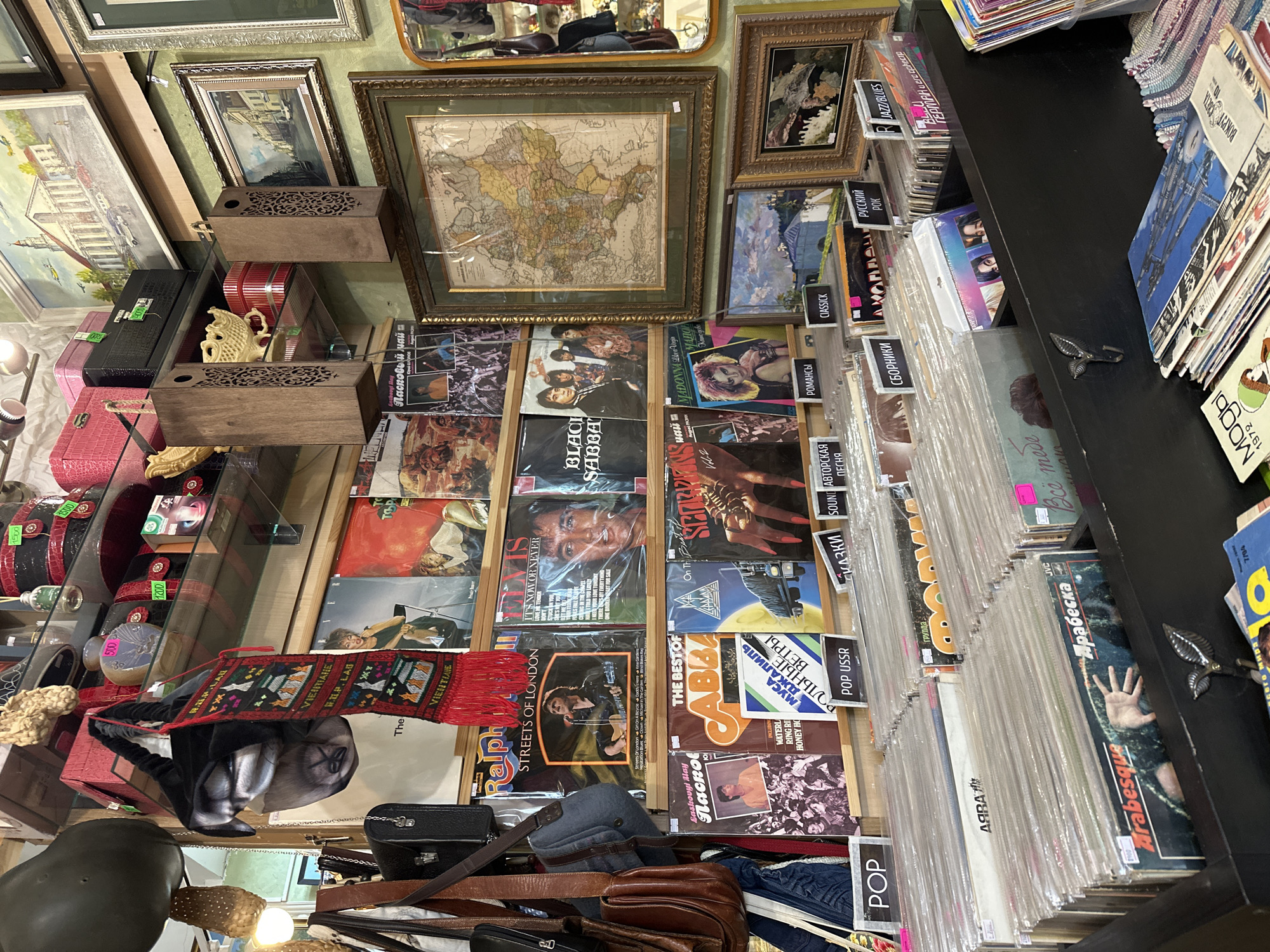The image shows an indoor space filled with various items. Frames of different sizes hang on the wall, including one with a map. Below the frames are shelves filled with vinyl records. The record covers feature a variety of designs and titles, with price labels visible. Other items are displayed in a glass-enclosed area, including purses, boxes, and other decorative objects. A scarf with the word "VIENTIANE" is draped on a purse. A light fixture is visible in the lower left corner. Based on the presence of Cyrillic labels and pricing, the location is likely a store in the city of Kazan, Russia, where such records are sold.