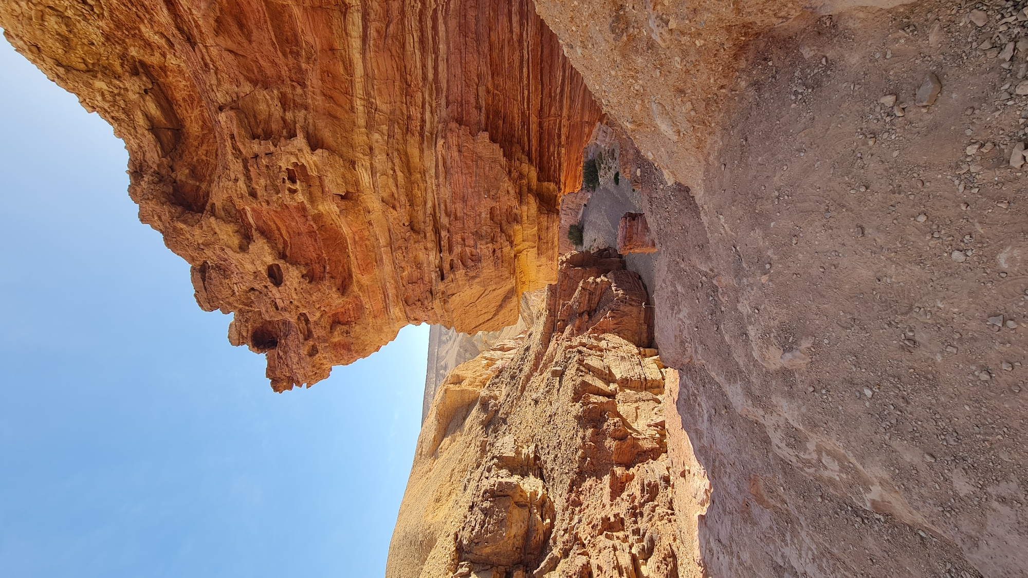 The image, taken at an unusual angle, presents a scene of rugged natural beauty in the Hevel Eilot Regional Council, Israel. The dominant feature is a massive, overhanging rock formation, characterized by its layered, reddish-brown strata, with some areas having holes or being porous, giving it a textured appearance. Below the overhanging rock is another rock formation and some soil. The angle of the photo makes it difficult to identify exactly what is going on, but you can see a road below the rock formations. A clear, blue sky forms the backdrop in the top left corner of the frame. The overall setting evokes a sense of a desert landscape.