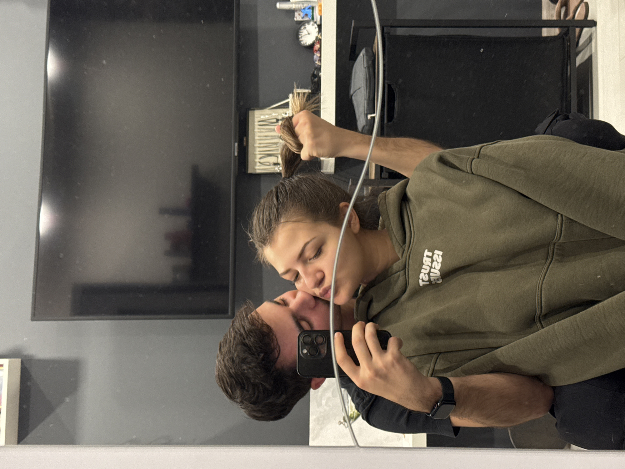 A couple takes a selfie in front of a mirror. The woman, wearing an olive green hoodie with the words "TRUST ISSUE" printed on it, is pulling up her hair in a ponytail with her right hand. The man, wearing a dark shirt, holds a black smartphone to take the picture, their faces nearly touching as if about to kiss. The environment shows a room with a large flat-screen TV on the wall. A shelf behind them holds various items including a clock, jewelry box, and a black bag. There is no clear indication of the location in the city of Sofia, Bulgaria.