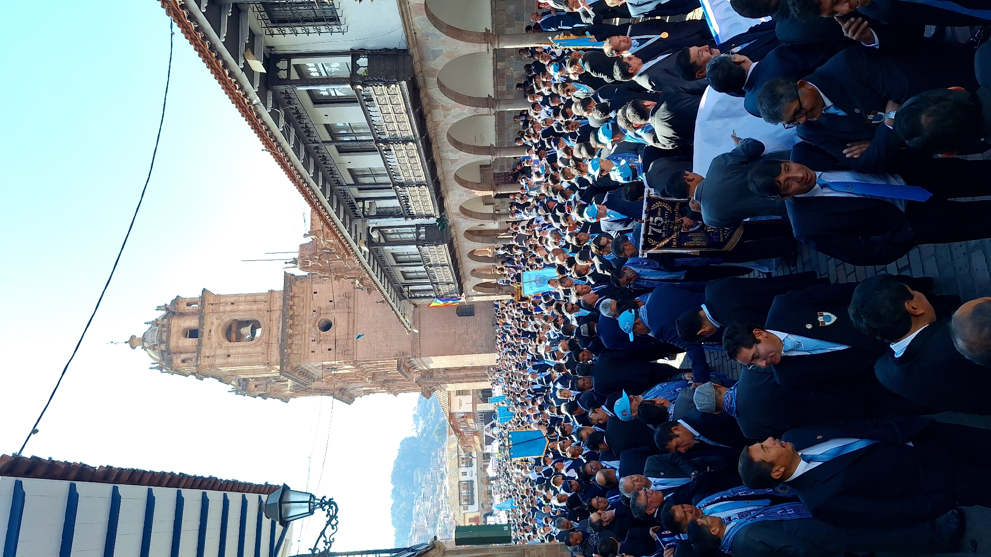 This is a high-angle, wide shot photograph captured on a bright, sunny day in Cusco, Peru. The scene depicts a large gathering of men, predominantly dressed in dark suits and light blue accessories like ties and scarves, participating in a procession or parade. Many are wearing blue caps. They are standing in a cobblestone plaza, bordered by colonial-era buildings with arched walkways and terracotta tiled roofs.
Dominating the background is a prominent bell tower of a historic cathedral, likely the Cusco Cathedral, a significant landmark in the Plaza de Armas. The architecture is distinctly Spanish colonial, with ornate balconies and windows. Several large blue banners are visible amongst the crowd, some bearing text that is difficult to decipher from this distance. In the foreground, a banner reads "75 AÑOS DE DILIGENTE G." and mentions "INCA GARCILASO DE LA VEGA", indicating a possible anniversary celebration or commemoration related to an institution or historical figure. The crowd extends far into the distance, filling the plaza and spilling into the surrounding streets. The presence of the colonial architecture and the cathedral tower strongly places the event in Cusco's Plaza de Armas.
