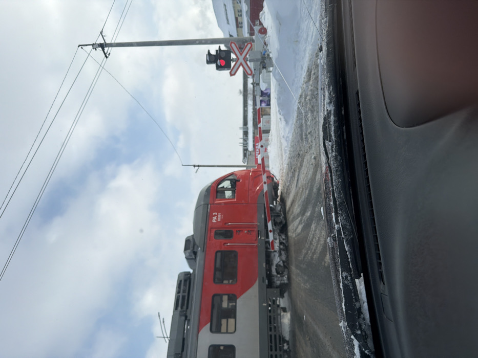 A red and grey train approaches a railroad crossing, where the gate is down and the signal lights are flashing red. Snow covers the ground on either side of the tracks. The image is taken from inside a car, as evidenced by the dashboard and window frame visible on the right side. Overhead electrical lines are also visible.
