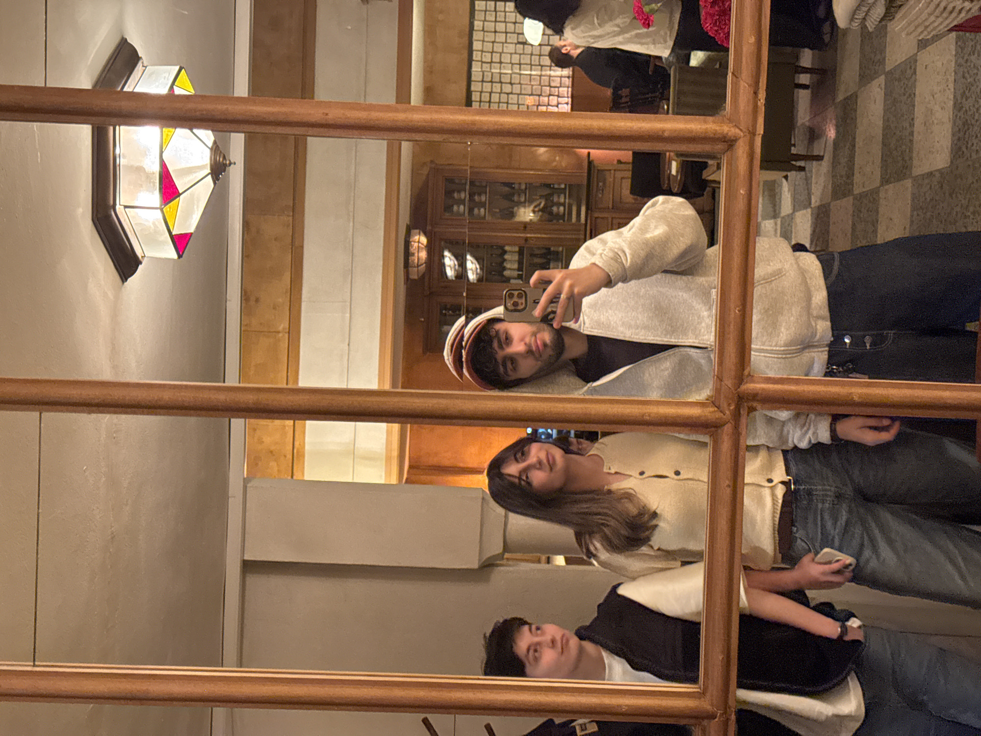 A medium shot shows a mirror's reflection of three individuals, likely in a restaurant or bar. The reflection shows a man in a light-colored hoodie and red cap taking a selfie with his phone. Behind him, a woman in a light-colored sweater holds her own phone. Below her is another person in a dark jacket. In the background is a tiled floor and a wooden cabinet containing bottles. A decorative lamp is visible to the side. While the location is possibly in Yerevan, Armenia, as stated in the instructions, there are no clear indicators to confirm this.