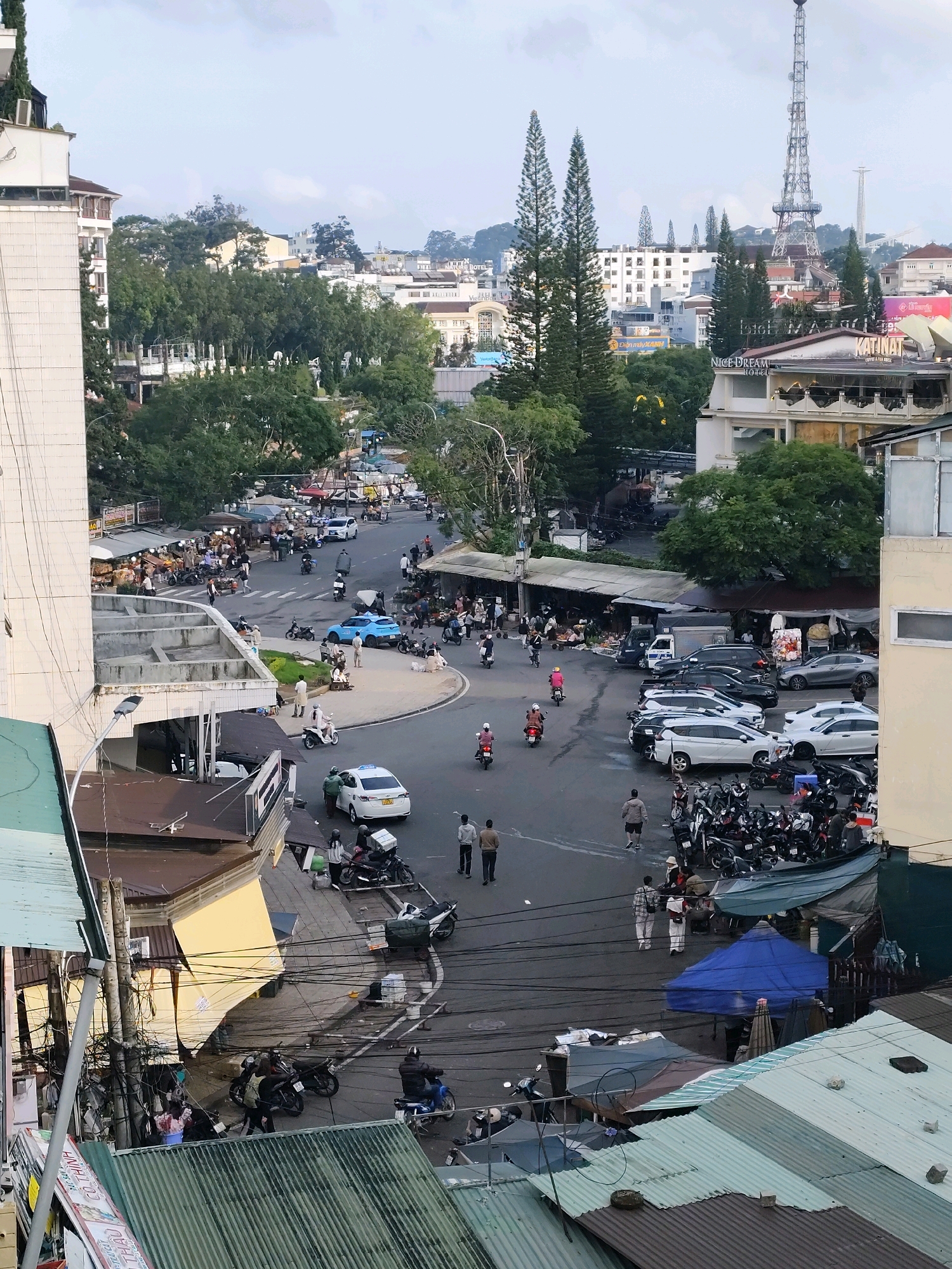This is an aerial, daytime, wide-angle shot of a busy intersection and surrounding cityscape in Da Lat, Vietnam. The foreground features several corrugated metal roofs. A white car with a license plate is parked on the right side of the intersection, adjacent to a building with a light-colored facade. A group of motorcycles is parked on the road and sidewalk.
In the middle ground, the intersection is bustling with traffic, including motorcycles and cars. Several people are walking and standing on the sidewalks and in the street. Some are carrying bags or pushing carts, suggesting a market or commercial area. Various stalls with awnings are set up along the sides of the road. There are several buildings with a mix of architectural styles, including a tall white building on the left.
In the background, the cityscape extends with more buildings and trees. A prominent telecommunications tower, resembling the Eiffel Tower, stands tall in the distance, along with other structures. The sky is overcast with clouds. The overall scene depicts a lively urban environment with a mix of residential and commercial activity, characteristic of a Vietnamese city.