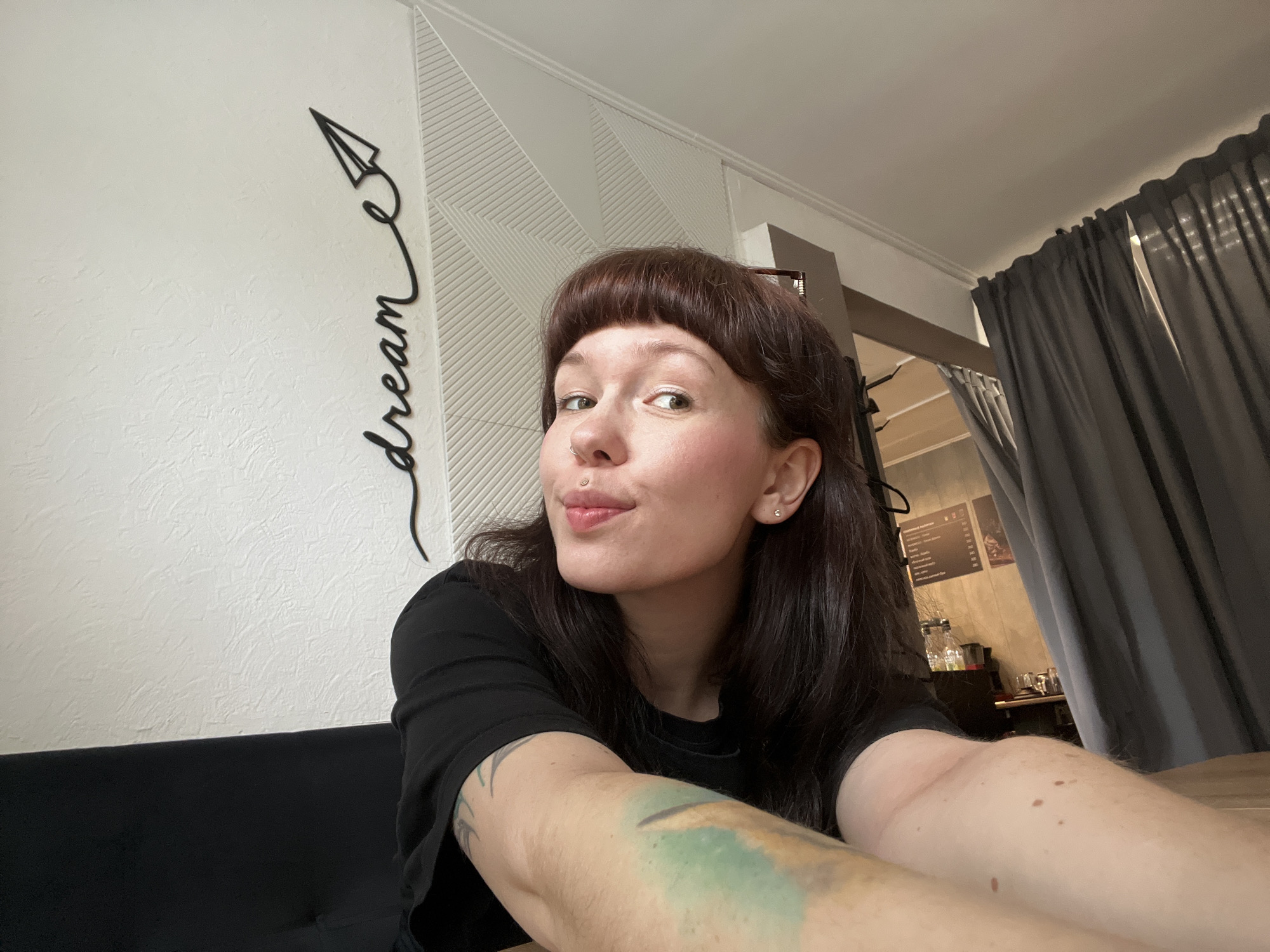 The image is a selfie of a woman with brown hair and short bangs. She is wearing a black t-shirt. The location appears to be indoors, possibly a cafe or restaurant, as suggested by the decor and the presence of curtains. Behind the woman is a wall with a "dream" sign and a geometric pattern. A door or entryway is visible in the background. The woman has a nose piercing and a lip piercing, and a tattoo is visible on her arm. It is not possible to confirm the location in Chaikovsky, Russia from the given visual information.