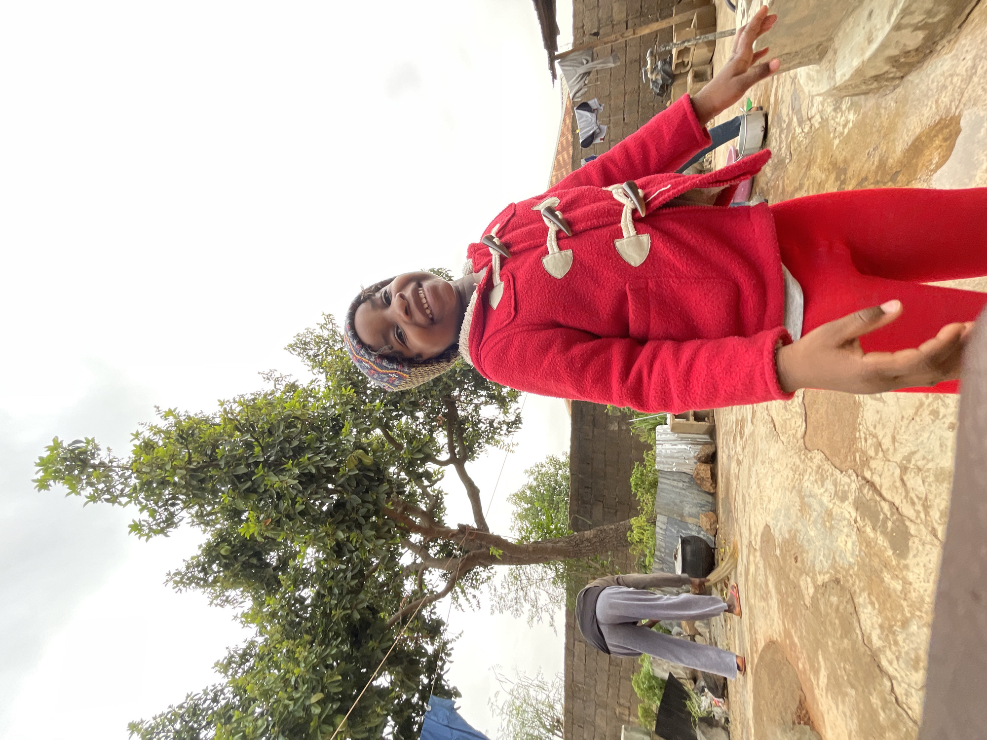 In Jos, Nigeria, a young girl stands outdoors, smiling and gesturing with her hands. She wears a red coat, red leggings, and a patterned knit hat. Behind her is a tree with green leaves. A person in gray clothing is sweeping the ground near a corrugated metal fence. Clothes hang on a line in the background. The ground appears to be a cracked, light-colored surface. The sky is overcast.