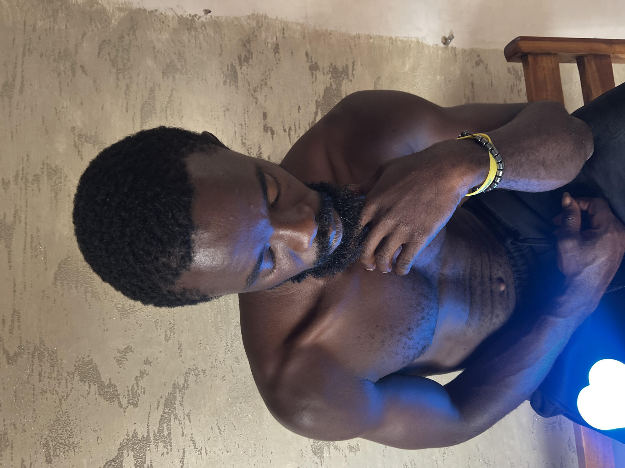 A shirtless Black man with a beard is pictured from the chest up, seated and looking down with his eyes closed. He is wearing black pants and a yellow and gray bracelet on his right wrist. His right hand is touching his chin. The background is a textured wall. There is a blue light illuminating the man's left shoulder and arm, and a white heart shape superimposed on his lower left side. The scene is set in a location within the city of Jos, Nigeria.