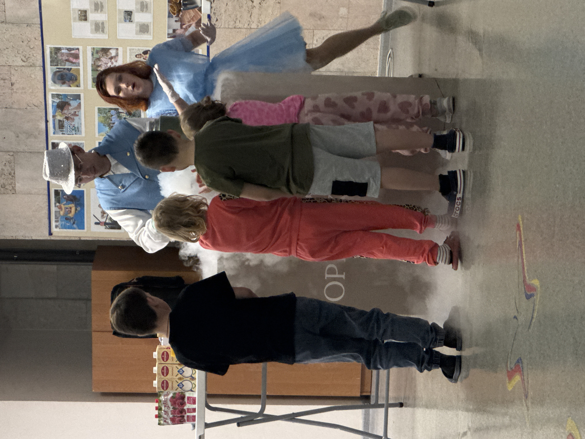 A magician in a light blue suit and white hat stands with his hand outstretched, seemingly conducting an invisible object. Next to him are five children lying on the floor in a straight line. The first child is wearing a blue tutu, the second is wearing pink pajamas with hearts, the third has on a green shirt and gray shorts, the fourth wears a red jumpsuit, and the fifth has on a black t-shirt and jeans. Between the magician and the children is a wisp of white smoke.
To the left is a table with juice boxes and juice pouches. Behind the magician are pictures displayed on the wall. The word "OP" is visible on the floor, and a colorful star is drawn on the floor near the bottom of the image.
The location is the city of Yevpatoriya, Ukraine.