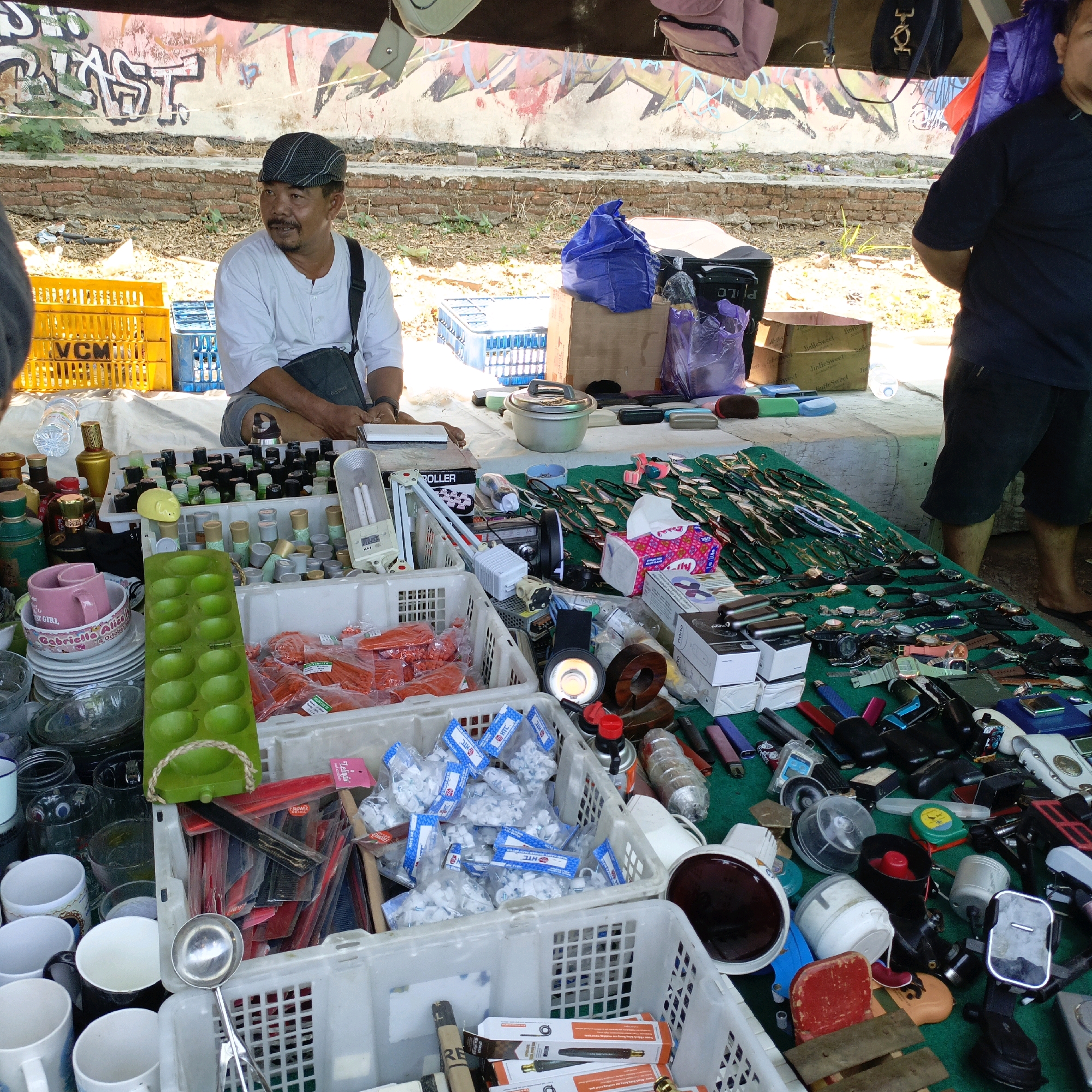 A street-side market stall is set up under a tent, displaying a variety of goods. A man wearing a cap and a white shirt sits behind the stall, looking to his right. Several plastic crates filled with items are visible, including yellow and blue crates in the background and white crates in the foreground containing packaged goods, red-orange plastic items, and what appear to be small white fasteners. On a green cloth covering a table, numerous items are laid out, including rows of watches, sunglasses, and lighters. Various bottles, cups, bowls, and kitchen utensils are also present. The background features a wall with graffiti and a brick structure. The overall scene suggests a market in Ambarawa, Indonesia.