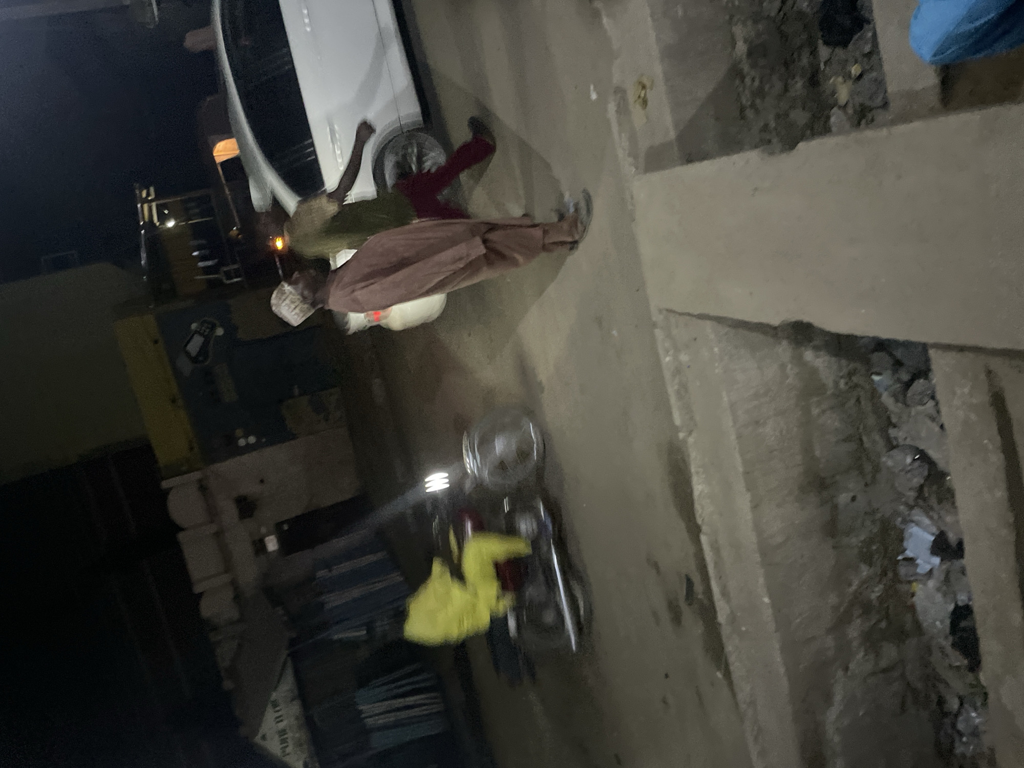 The image shows a nighttime street scene in an urban environment. A white car is parked near a sidewalk. Two individuals are visible near the car. One person is wearing a green shirt and red pants, while another individual, possibly elderly, is wearing a traditional robe and head covering. A motorcycle with a yellow cloth draped over it is parked on the side of the street. The scene is illuminated by streetlights and car headlights. There is trash visible near the curb. The specific location within the city is not determinable.