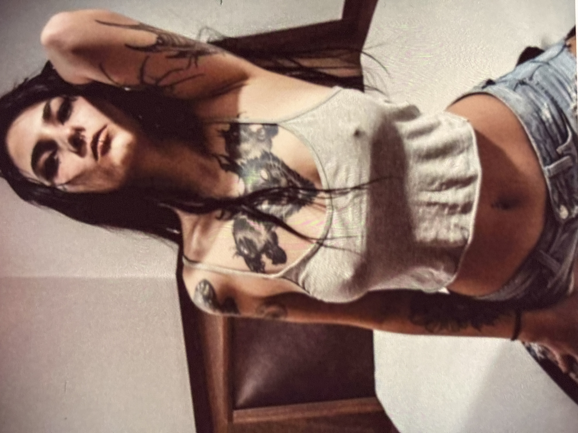 In an indoor setting, likely a bedroom, a woman with dark hair is depicted. She is wearing a light-colored tank top and denim shorts. Her body is adorned with tattoos on her arms and chest. The background includes a bed with a headboard and a wall. The lighting appears to be dim, creating shadows on the subject and the environment. The woman is posed looking towards the camera, with one arm raised.