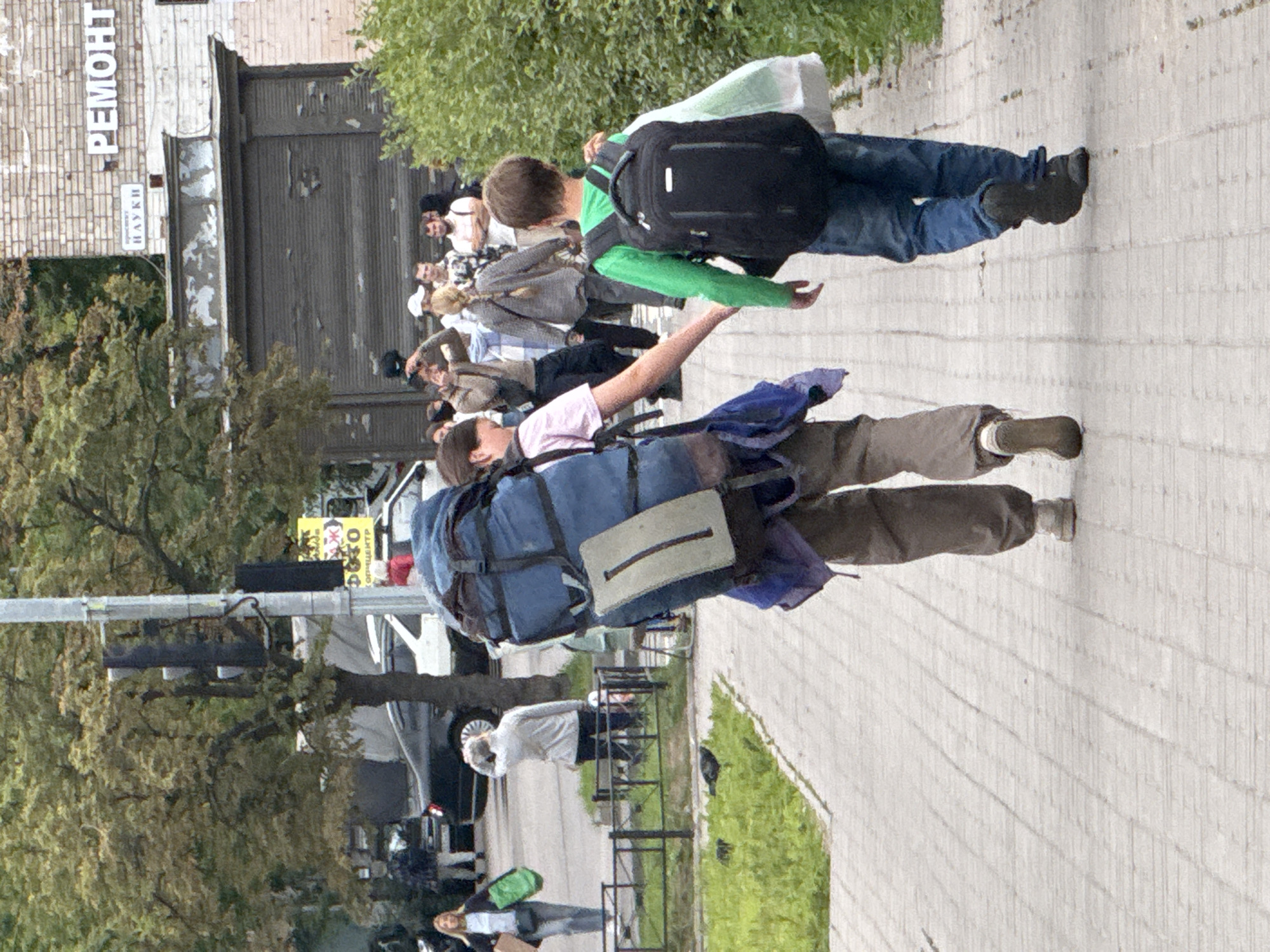 In Канонерец Снт, Russia, two individuals are walking on a paved sidewalk. The person on the left, with a large blue backpack, is wearing dark pants and white shoes. They are walking away from the camera. The person on the right, a younger individual, is wearing blue jeans and dark shoes, and also carries a black backpack and a white bag. They are walking in the same direction. Behind them, a group of people are visible in the background, some of whom appear to be looking at something or interacting. Trees and foliage are present along the side of the sidewalk, and a building with brickwork is visible in the distance. A traffic light is mounted on a pole to the left of the frame.