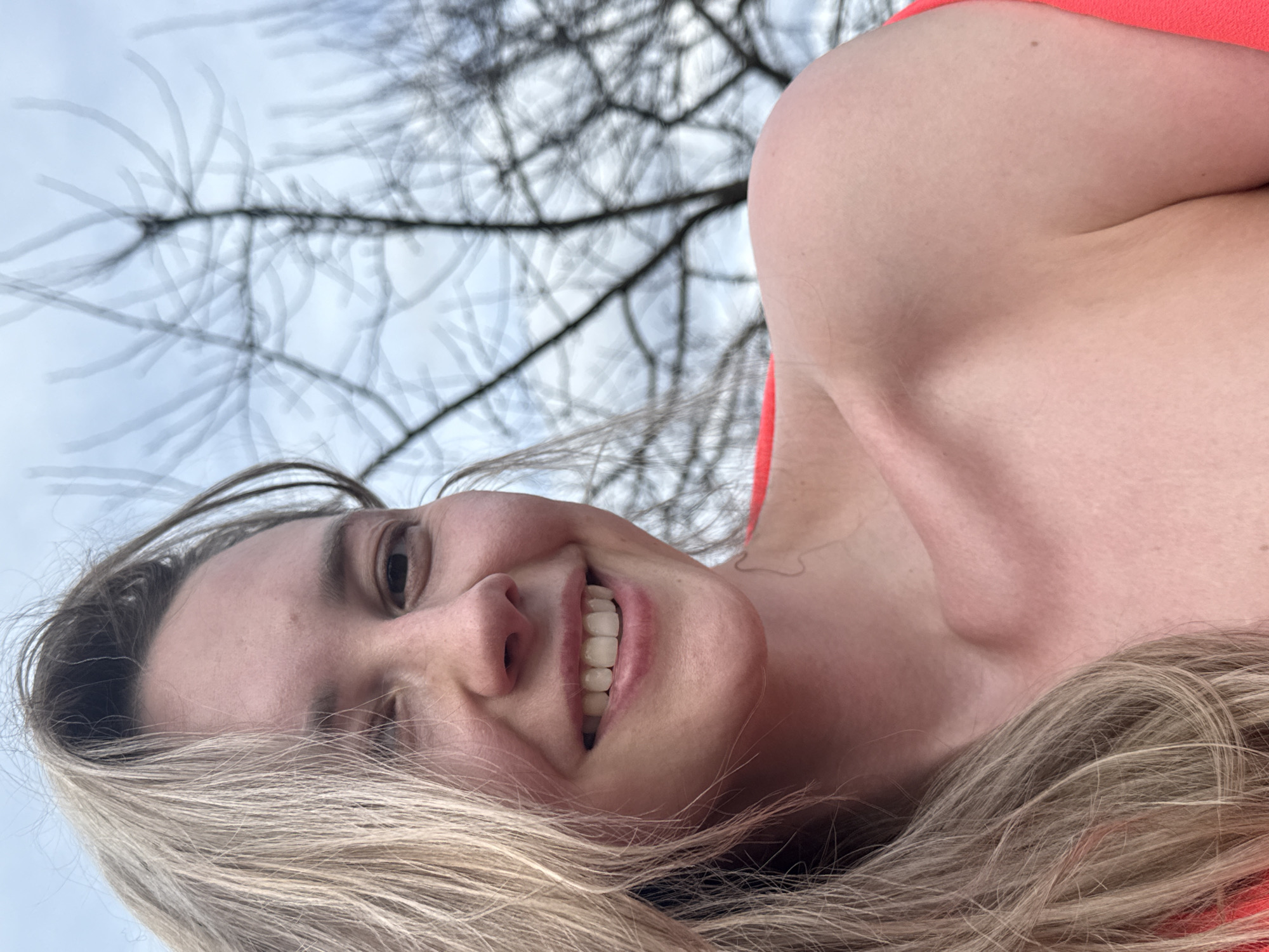 A close-up, low-angle selfie captures a smiling woman outdoors in Chaikovsky, Russia. The woman, with blonde hair flowing across the lower and left parts of the frame, is positioned in the foreground, looking up at the viewer with a wide, joyful smile, revealing her teeth. She appears to be wearing a red garment, visible on her shoulder and upper chest. The background features a light, overcast sky through which the dark, bare branches of trees are prominently visible. The tree branches exhibit a somewhat blurred or ethereal quality, suggesting either a creative photographic effect or movement against the pale sky. The overall scene conveys a casual, happy moment, likely taken during the day in a season where trees are bare, such as late autumn, winter, or early spring. No text is visible in the image.