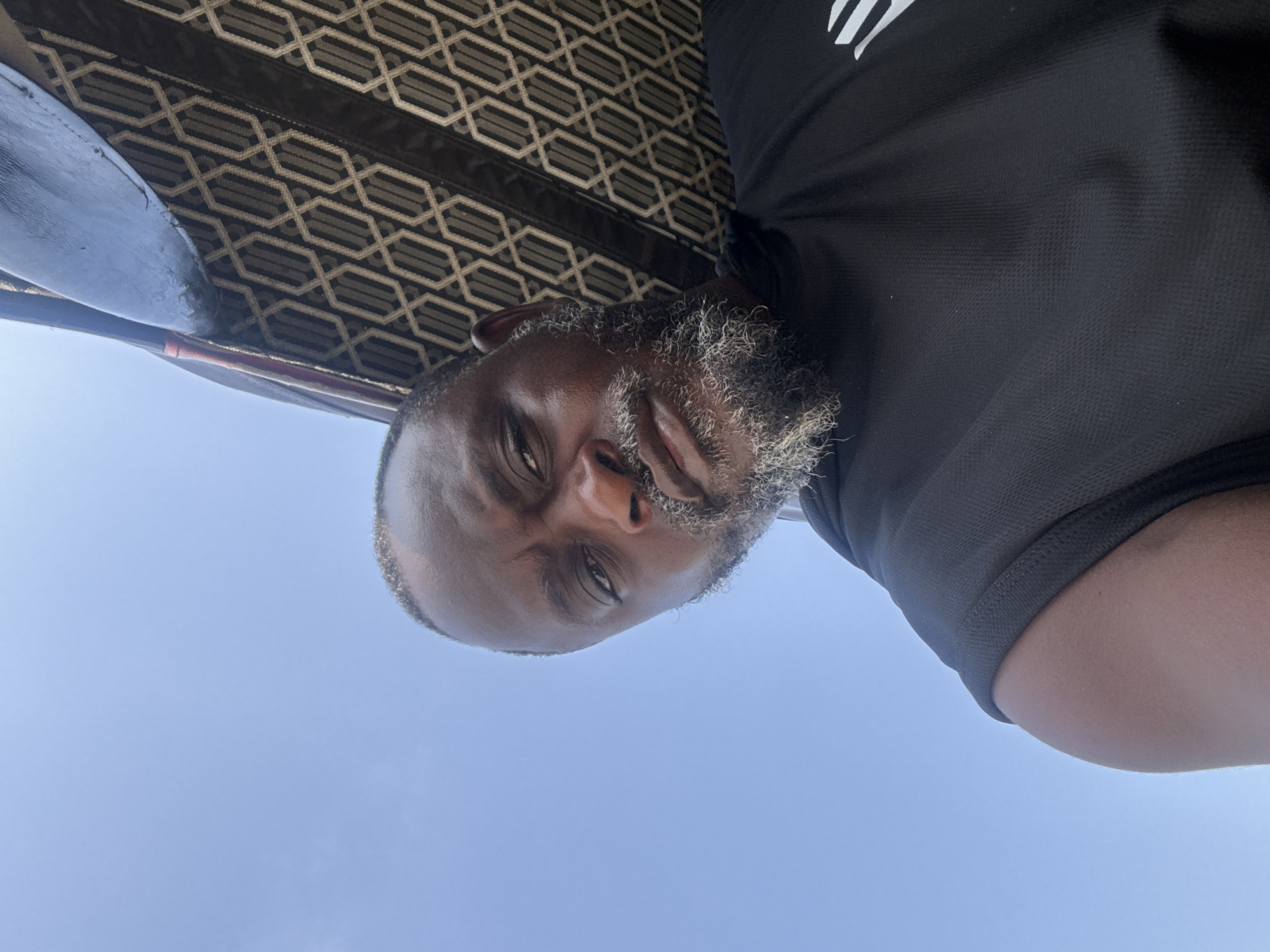 A selfie-style image shows a middle-aged black man with a salt-and-pepper beard. He is wearing a black shirt with a partial white logo on the shoulder. The background consists of a blue sky and a patterned ceiling. The location is unspecified but inferred to be Ado, Nigeria.