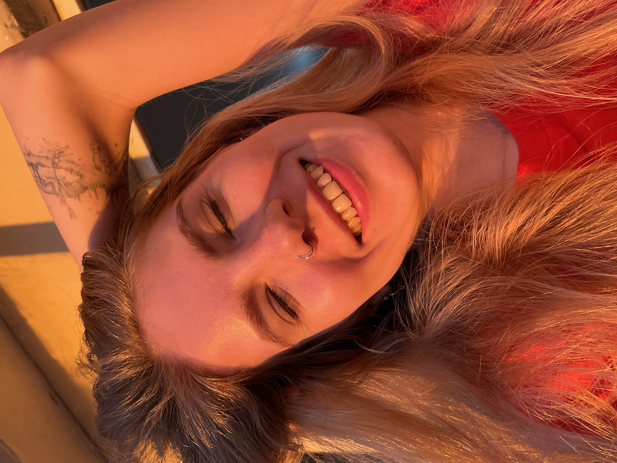 A young woman with blonde hair and a nose ring smiles with her eyes closed, her right arm raised and behind her head, revealing a tattoo on her upper arm. The image is bathed in warm, golden-orange light, suggesting it was taken during sunset or sunrise. She is wearing a red top. The background shows an illuminated, textured wall and a dark area. The scene appears to be outdoors. There is no discernible information within the image to indicate a specific location in Izhevsk, Russia.
