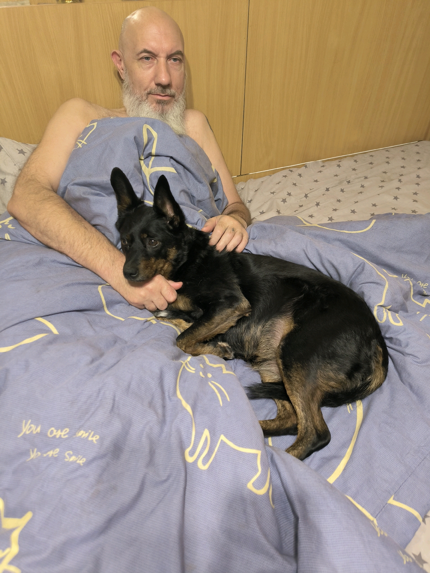 A bald, fair-skinned man with a grey beard is lying in bed, covered by a blue comforter with a yellow line drawing of a cat and text that reads "You are smile, You are smile." A medium-sized black and brown dog is lying on the comforter, partially on top of the man's arm. The man's left arm is visible, and his hand is resting on the dog's body. The background shows a wooden headboard. The scene appears to be indoors, likely in a bedroom. The location is unknown.