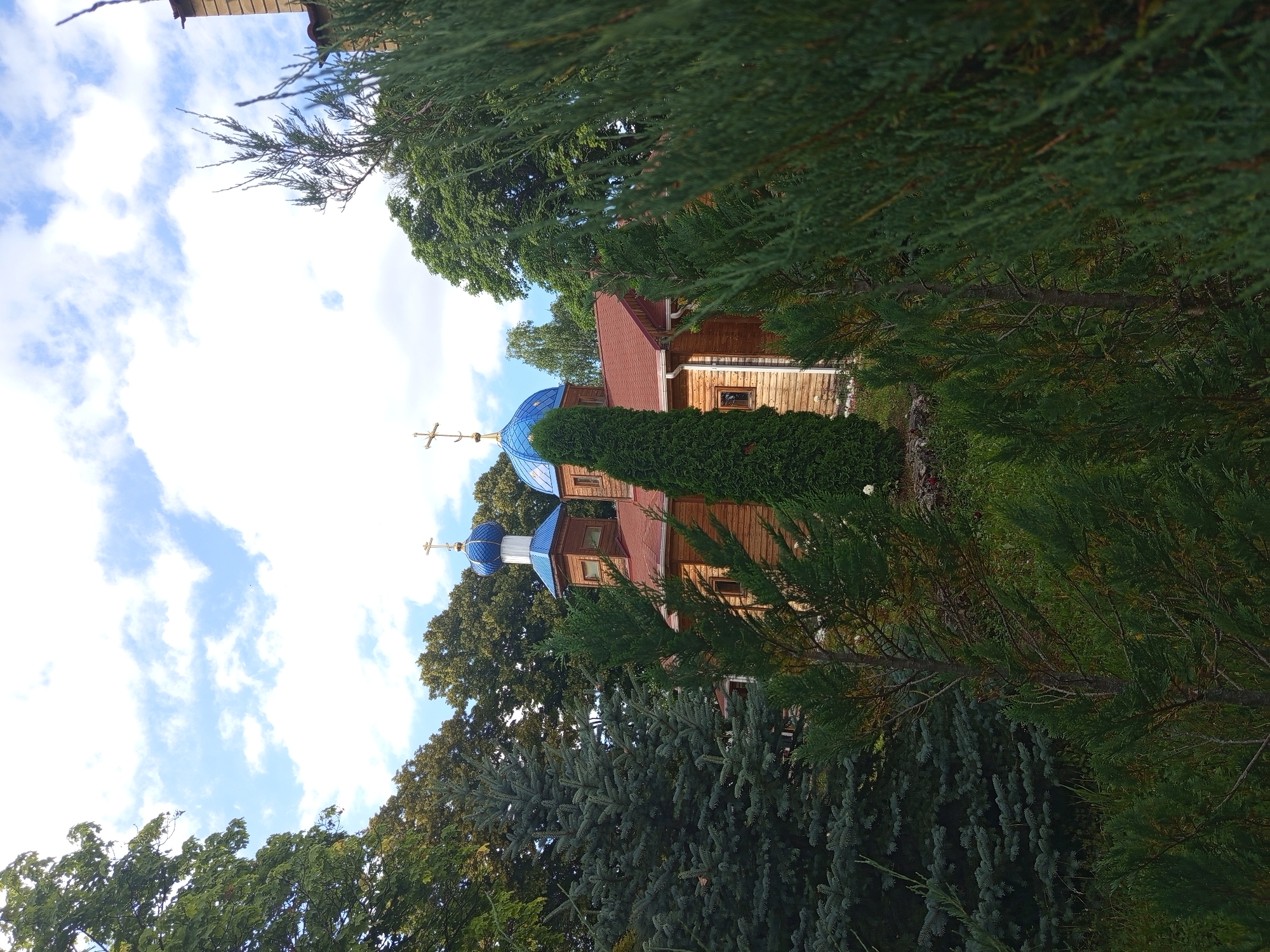 A wooden church with blue domes and golden crosses stands amidst lush greenery. The church has a red roof and visible windows. Tall evergreen trees and deciduous trees surround the structure. The sky is partly cloudy with blue patches. The scene appears to be located in a serene, possibly rural or park-like setting within Giaginskaya, Russia. There are no people visible in the image.