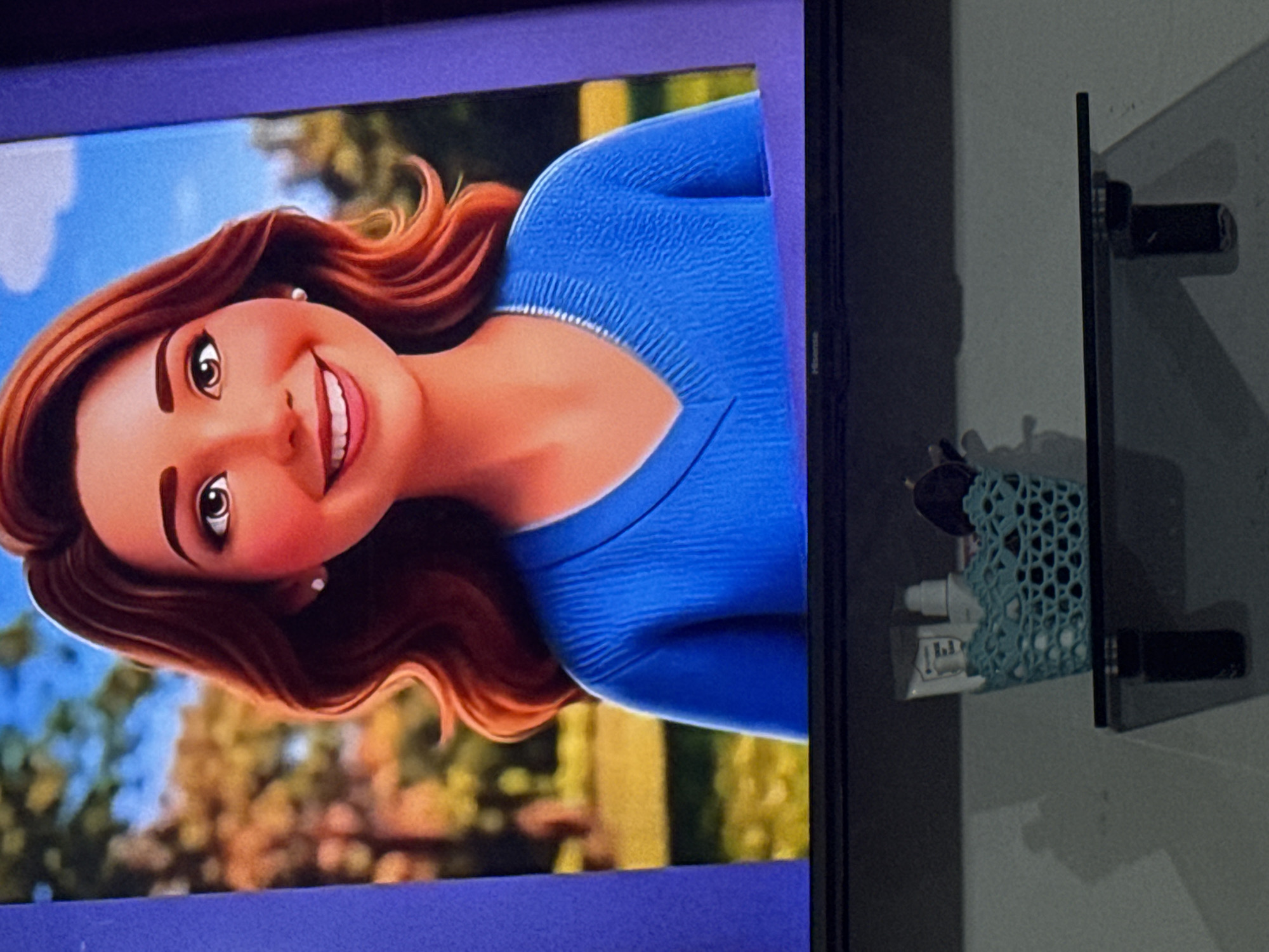 The image shows a close-up of a digital rendering of a woman with reddish-brown hair and brown eyes, wearing a blue v-neck sweater. Her expression is a wide smile, and she is wearing a pearl earring. The background behind her appears to be a park scene with trees and a blue sky. Below the digital display, a glass shelf is visible, on which sits a small, light blue woven basket containing toiletries and a dark object, possibly sunglasses. The shelf is attached to a wall, and there are shadows cast on the wall and the surface below. The location is identified as Chardara, Uzbekistan.