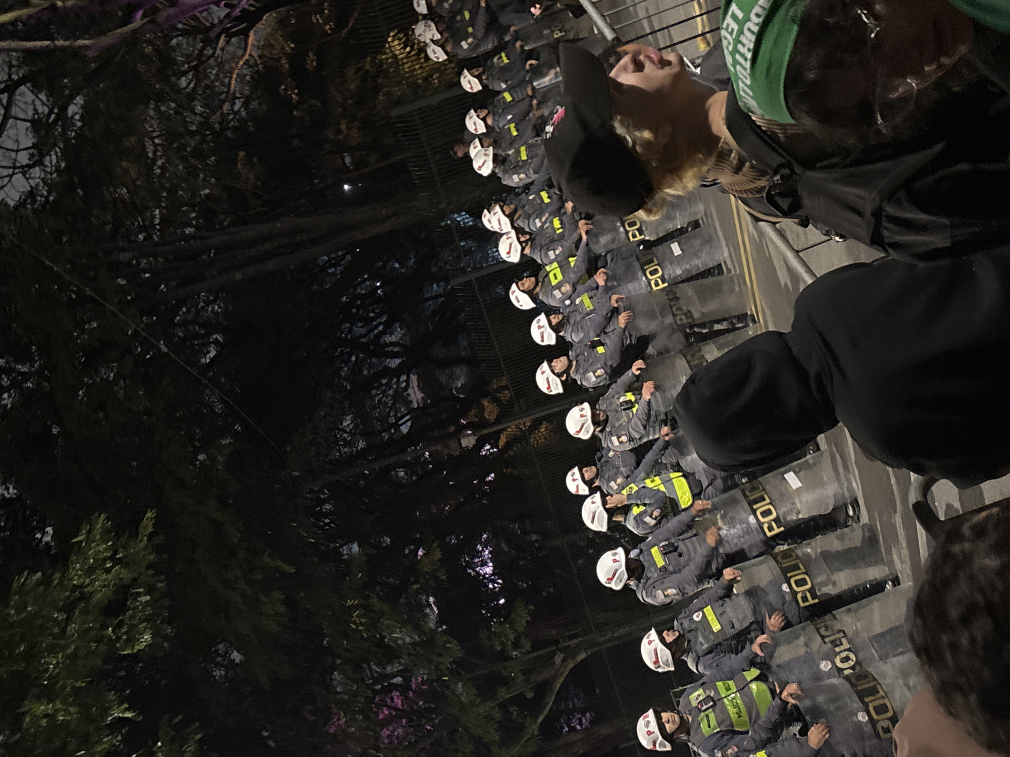 A nighttime image shows a row of riot police standing behind shields that read "POLICE," facing a crowd. The police are wearing helmets and vests. Trees are visible on the left, and people are in the foreground. The location is potentially in a city setting, possibly São Paulo, Brazil.