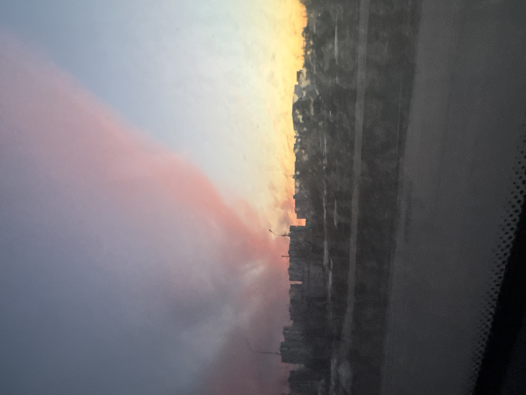 The image captures a view from inside a vehicle in Berezniki, Russia. Through the windshield, a cityscape is visible during a sunset. The sky displays a gradient of colors, transitioning from deep blues and purples on the left to orange and yellow hues above the buildings on the right. The urban landscape consists of a series of buildings that appear silhouetted against the vibrant sky. The interior of the vehicle, including the edge of the windshield with visible detail, frames the scene.