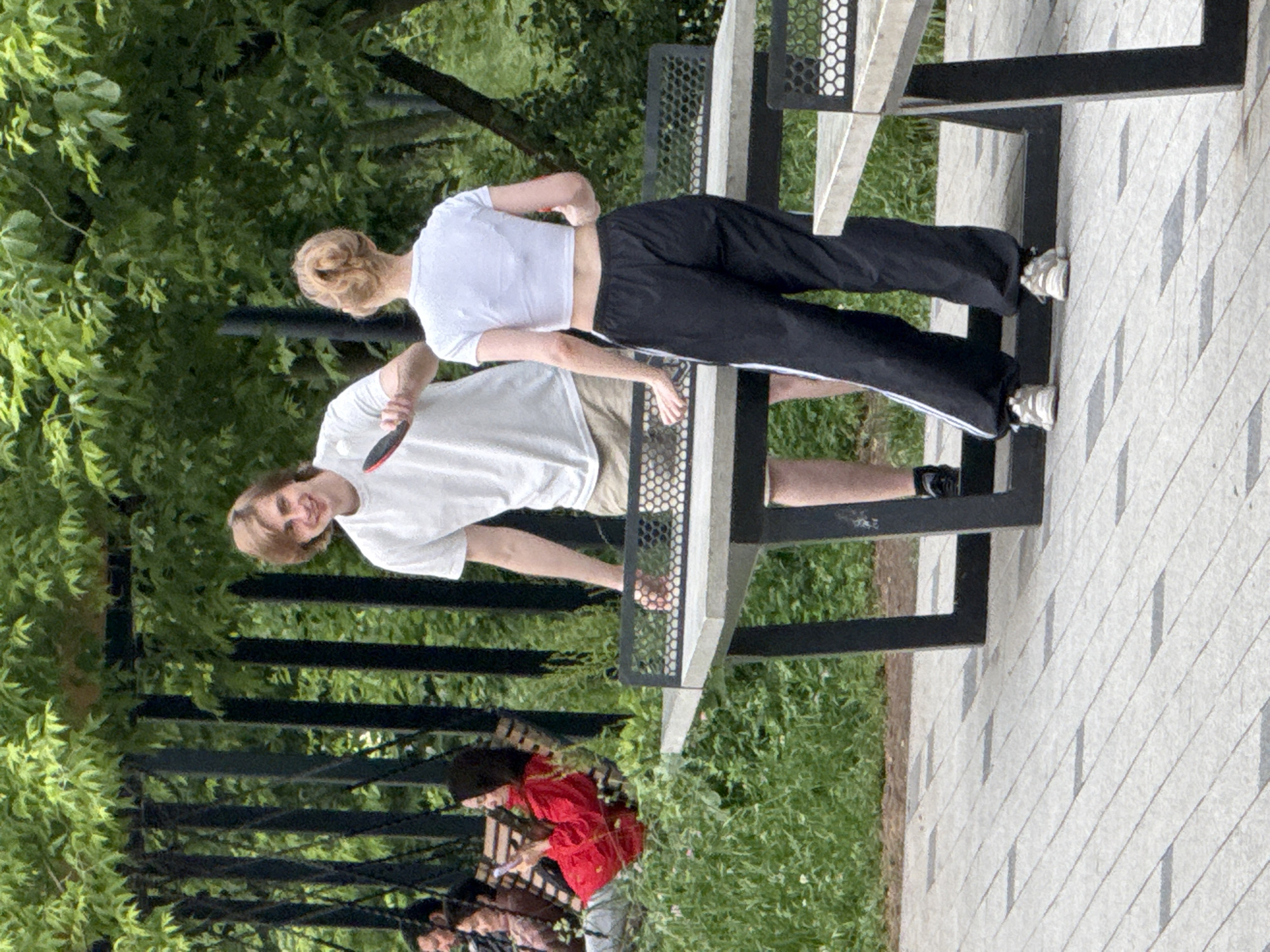 A young man and woman are playing ping pong outdoors. The man, wearing a white t-shirt and khaki shorts, is holding a ping pong paddle and looking at the camera with a smile. The woman, wearing a white crop top and black track pants, is facing away from the camera. They are standing on a paved area next to a ping pong table. In the background, two other individuals are seated on a bench swing, with one of them looking at their phone. The scene is surrounded by lush green foliage and trees, suggesting a park or outdoor recreational area.