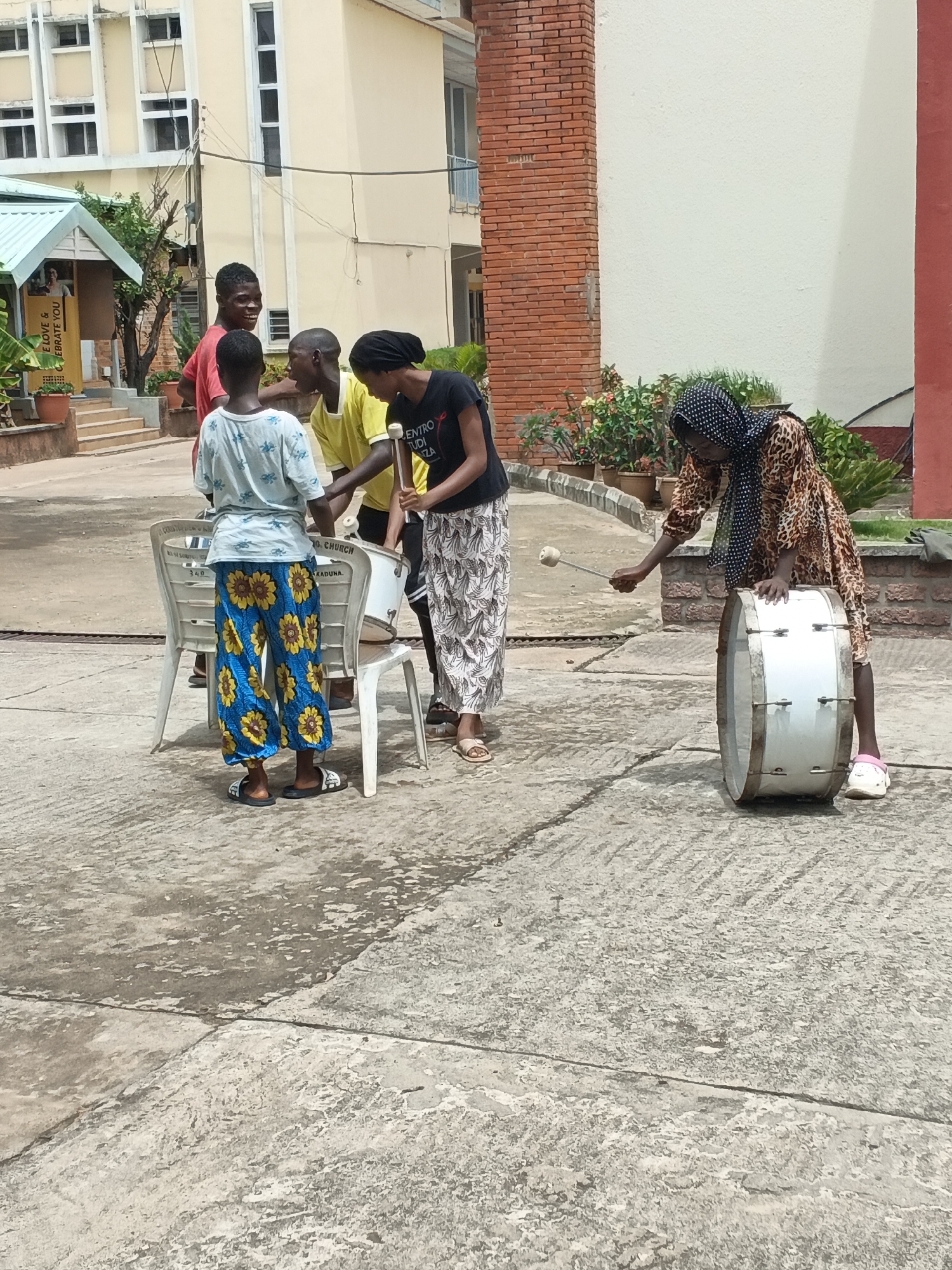 The image depicts a group of five people gathered outdoors in what appears to be a courtyard or open area in Unguwar Sarki, Nigeria. A woman wearing a leopard print dress and headscarf is striking a large white drum with a drumstick. Four other individuals are gathered around a smaller drum placed on top of two plastic chairs. The person closest to the camera is wearing blue and yellow flower-printed pants, and the others are wearing a red shirt, a yellow shirt, and a black shirt respectively. The surrounding environment includes building exteriors, potted plants, and tiled surfaces.