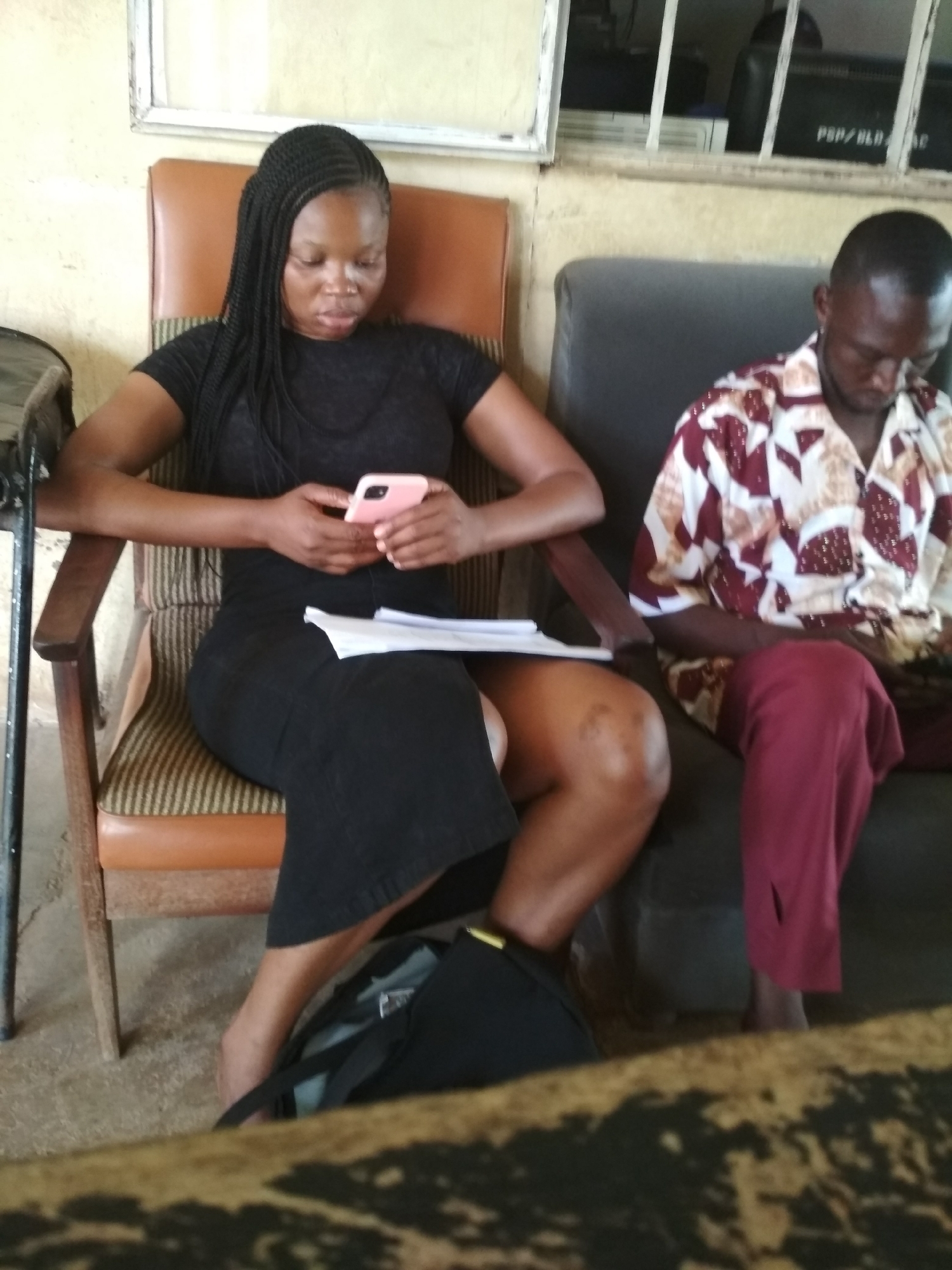 In Bisichi, Nigeria, a woman with braided hair sits on a patterned chair, holding a pink smartphone and a document. She wears a black dress. Beside her, a man in a patterned shirt and maroon trousers sits on a dark couch, looking down. A dark bag is on the floor near the woman's feet. The environment appears to be an indoor waiting area with light-colored walls and a barred window in the background.
