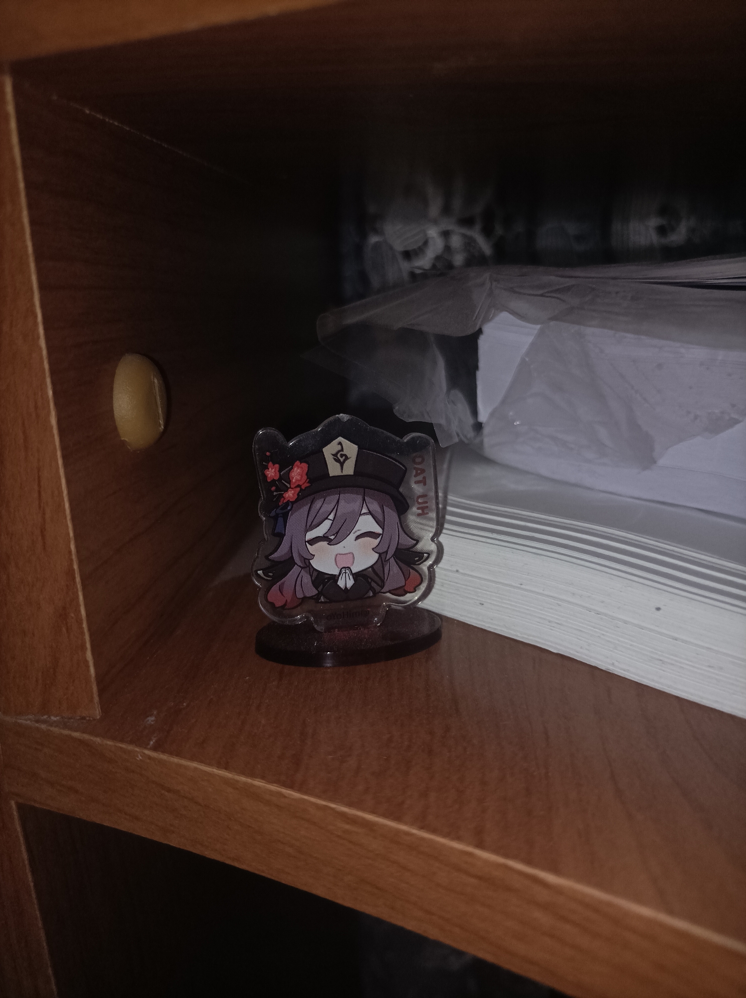An anime-style acrylic standee of the character Hu Tao from Genshin Impact is displayed inside a dark wooden shelf or cabinet compartment. The character on the standee has purple hair, wears a black hat adorned with red flowers, and a dark outfit. She is depicted with a wide, happy smile and her hands clasped together, suggesting a cheerful or polite gesture. The standee is placed on a dark circular base.
The overall scene is dimly lit, casting shadows within the compartment. To the left of the standee, a light-colored, oval-shaped object is affixed to the wooden back wall, possibly a sticker or a piece of hardware. A thin strand resembling a cobweb or dust line is visible on the upper left interior corner of the shelf.
In the background, behind and to the right of the standee, several stacks of white papers or documents, partially wrapped in clear plastic, are visible. The wooden surfaces of the shelf have a visible grain pattern.
Visible text on the standee includes "OAT UH" along the side of the character's head, likely part of "HU TAO," and "OYO HU" (part of "miHoYo" and "Hu Tao") on the base, indicating the character's origin and developer. No activity is taking place; it is a still life image of a collectible item within a piece of furniture. The image was captured in Semyonov, Russia.