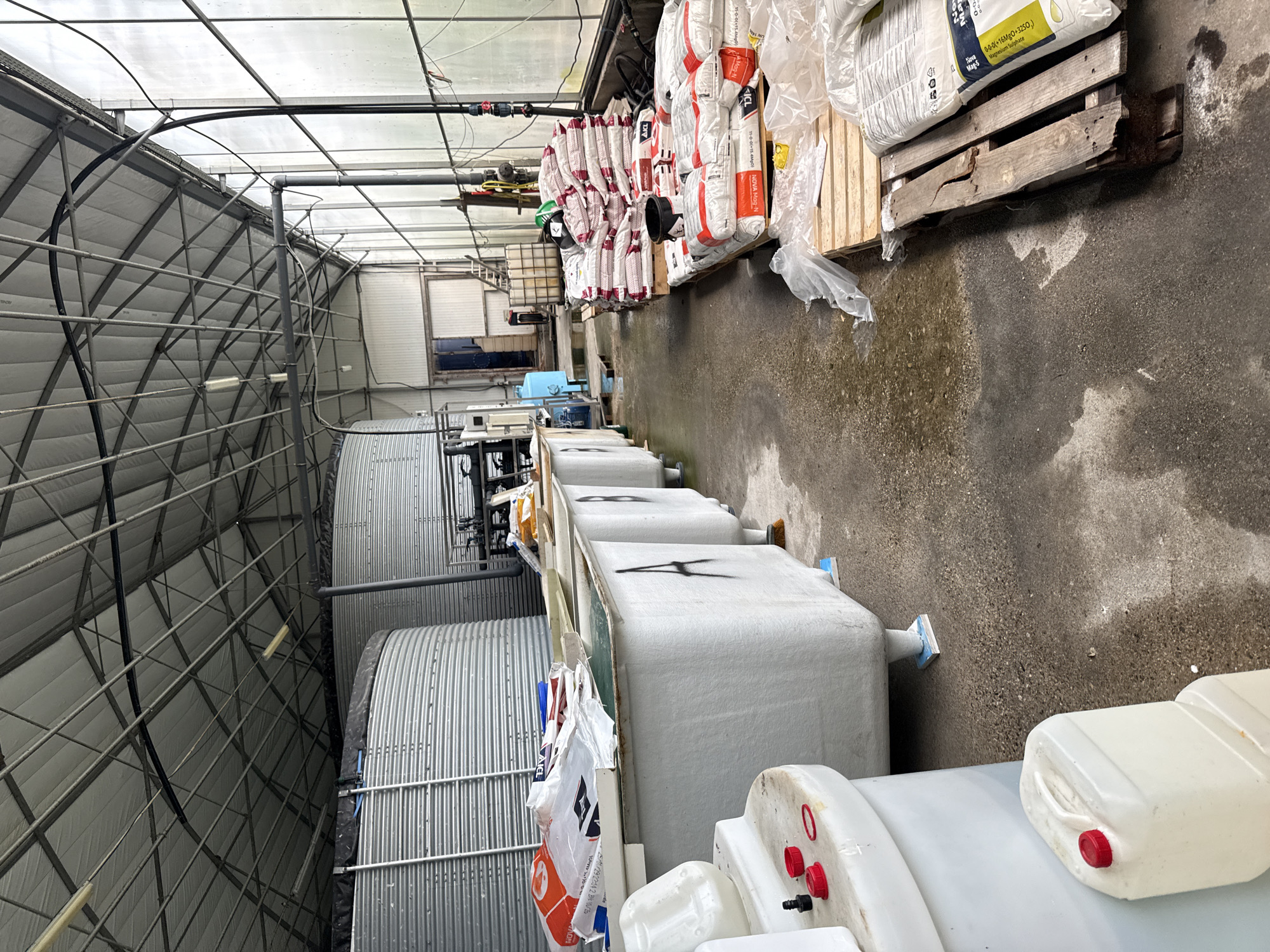 This image depicts the interior of what appears to be a greenhouse or agricultural facility. The most prominent features include rows of large, white tanks, potentially used for water storage or hydroponics. Adjacent to these tanks are tall, corrugated metal structures, possibly storage silos or supporting infrastructure.
The foreground shows an array of smaller, white containers, likely used for liquid storage, possibly chemicals or nutrients. In the background, there's a stack of large bags on wooden pallets, branded with labels like "ICL" and containing substances like Magnesium Sulphate. The bags appear to contain fertilizer and other agricultural chemicals. The environment is indoors, with a metal framework forming the greenhouse structure and a translucent roof providing natural light. The floor is concrete and shows signs of dampness or spills. There are no people visible in the frame. I cannot place this at a specific location in Stara Zagora, Bulgaria.