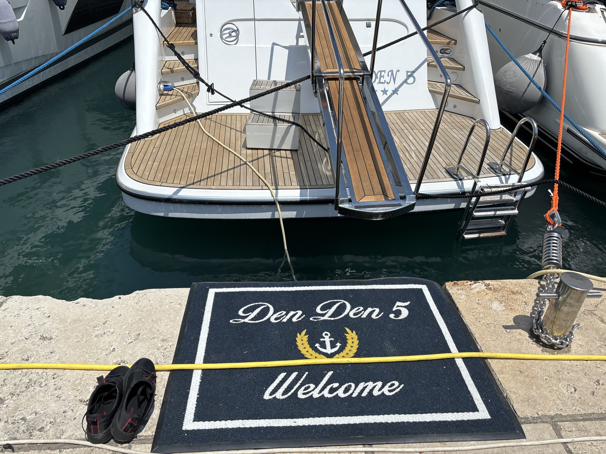 A pair of black athletic shoes with red accents are placed on a concrete pier next to a dark blue welcome mat that reads "Den Den 5 Welcome" with an anchor and laurel wreath emblem. The welcome mat is positioned in front of a yacht docked in clear blue water. The yacht has a wooden deck and a stainless steel gangplank. Ropes and fenders are visible, connecting the yacht to the dock and to another boat. The scene appears to be in a marina.