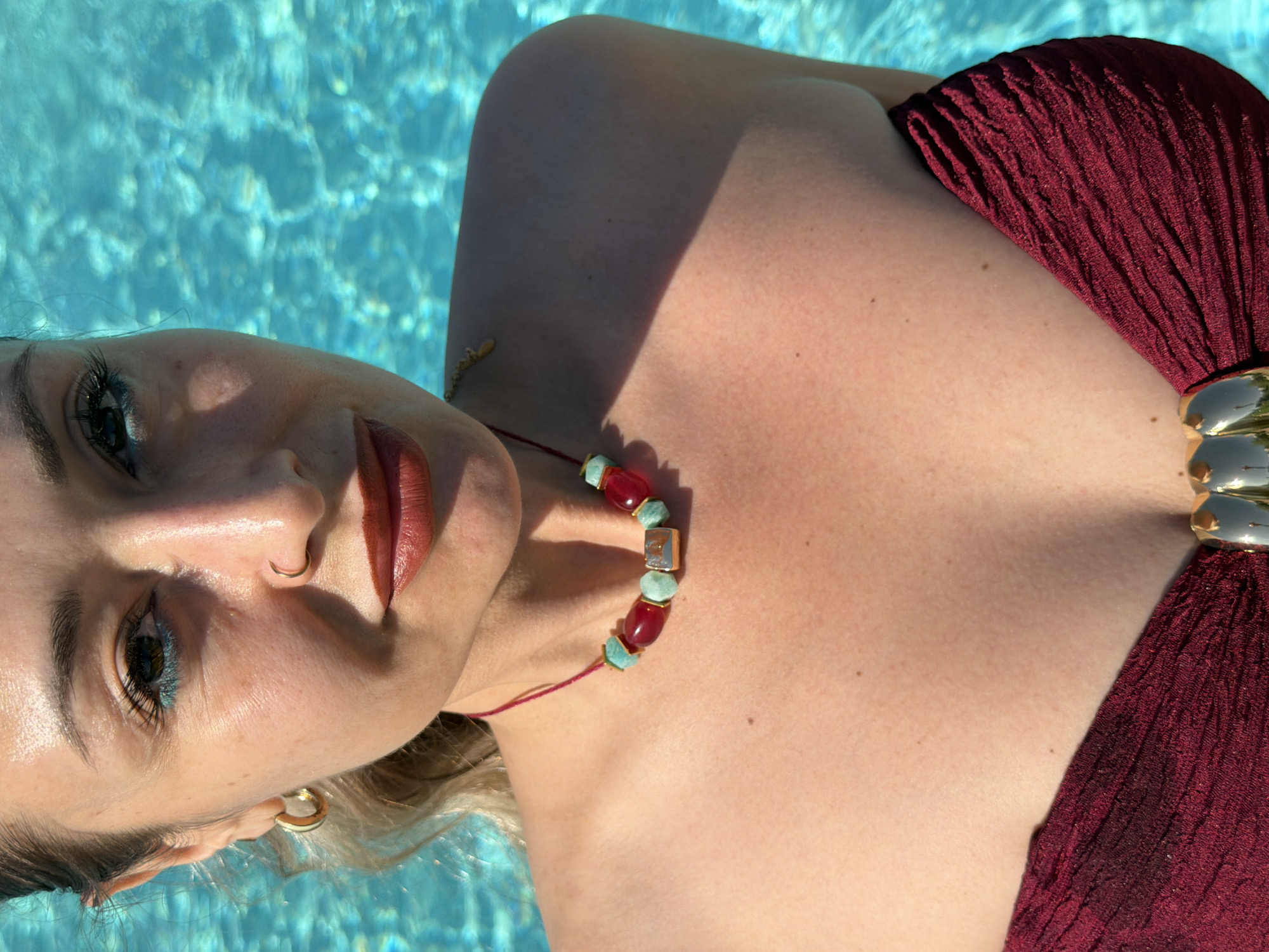 An overhead, rotated close-up shot captures a woman partially submerged in the clear, rippling blue water of what appears to be a swimming pool, likely in Krasnoyarsk, Russia. Her head is positioned to the left of the frame, tilted slightly upwards, with her body extending diagonally to the right. Bright, direct sunlight casts strong shadows across her face and décolletage, highlighting her fair skin.
The woman has brown eyes, accentuated by blue eyeliner and dark mascara, and wears reddish-brown lipstick. A small gold hoop nose ring adorns her left nostril, and a larger gold hoop earring is visible in her left ear. Her light-colored hair is mostly out of frame, but some strands are visible near her ear.
She wears a burgundy or deep red swimsuit top with a pleated or textured fabric, featuring a gold-toned, possibly metallic, ornamental clasp at the center of her chest. Around her neck is a distinctive necklace made of red spherical beads and light blue or turquoise-colored irregular, possibly square, beads, interspersed with smaller gold elements.
The water surface behind her is a vibrant light blue, reflecting the sun in a dazzling, textured pattern. The overall impression is one of relaxation in a sunny, aquatic environment. There is no visible text in the image.
