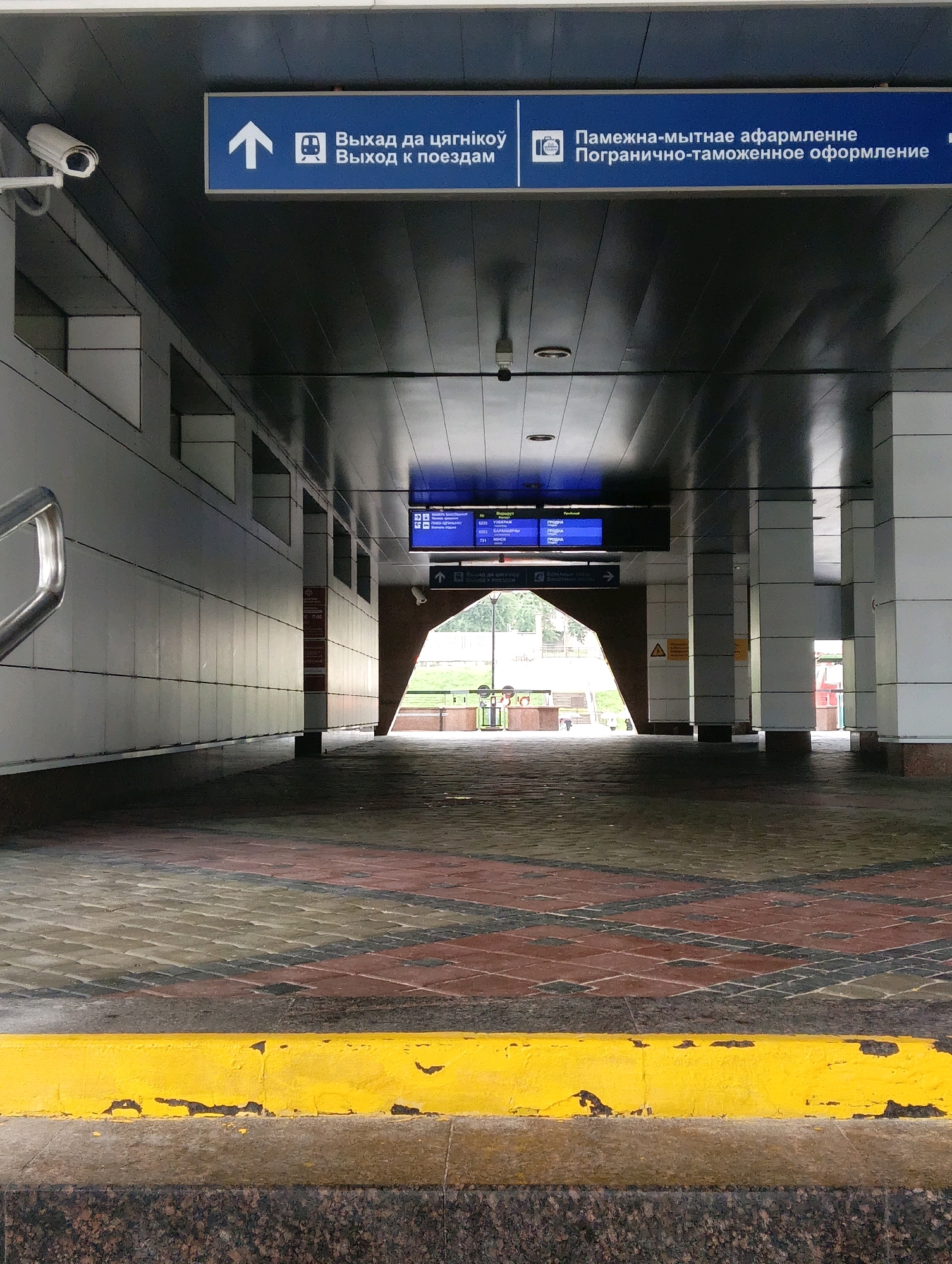 The image depicts an interior view of what appears to be a transportation hub, possibly a train station or bus terminal, in Hrodna, Belarus. The scene is captured from a low angle, looking down a covered walkway with a patterned tiled floor in shades of red, brown, and grey. A bright yellow barrier runs along the bottom of the frame, separating the walkway from a granite-like surface.
The ceiling of the walkway is dark and reflective, with various signs and lighting fixtures visible. Overhead, a blue sign with white text in what appears to be Cyrillic script indicates directions, with icons of an upward arrow and a train. Another similar blue sign further down the walkway displays more text and a camera icon. A security camera is mounted on the left wall, near the ceiling.
The walls of the walkway are light-colored and tiled, with a series of recessed rectangular openings. A metal handrail is visible on the far left. The walkway opens up to an area with natural light, suggesting an exit or a more open section of the terminal. In the distance, some greenery and structures are faintly visible. The overall atmosphere is functional and utilitarian, typical of public transport infrastructure. No people are clearly visible in the image.
