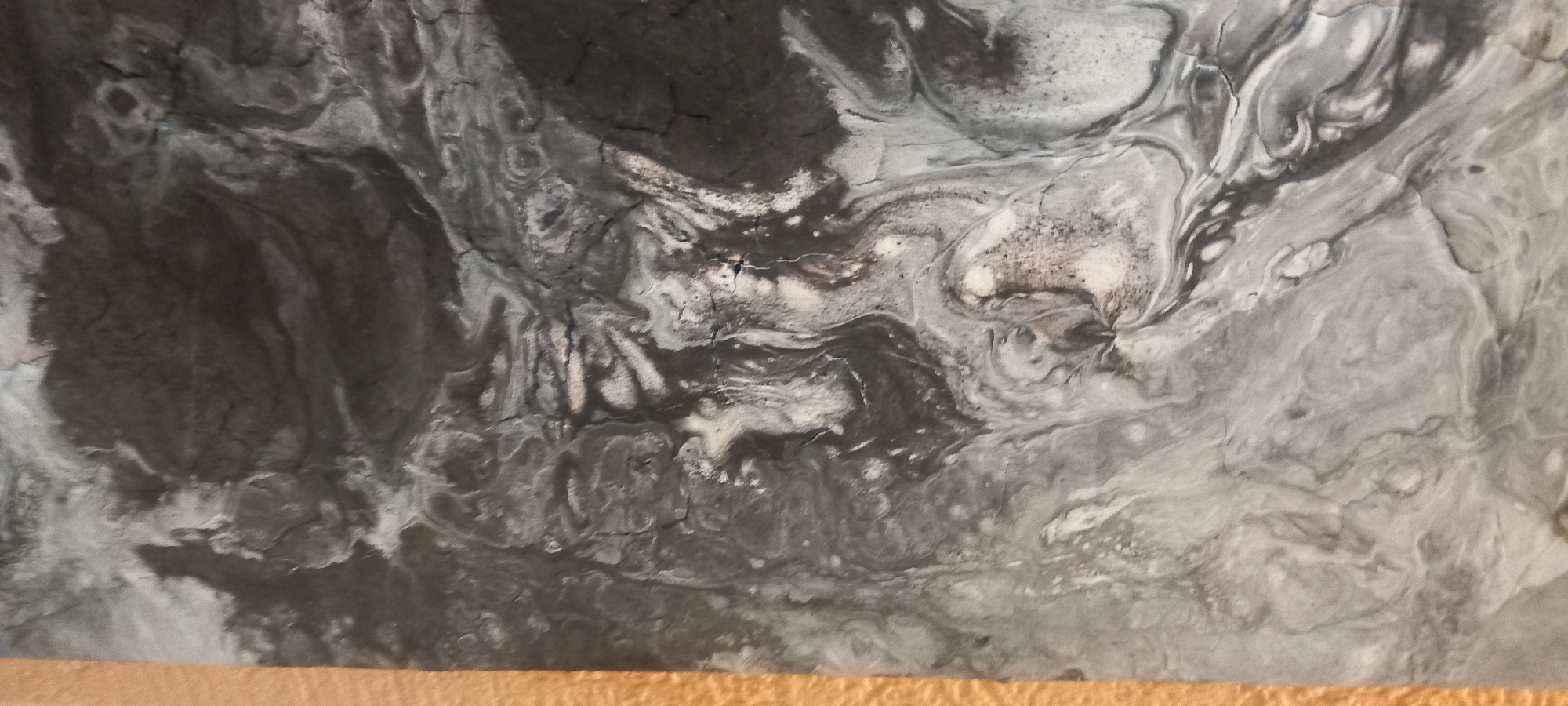 The image is a close-up, vertically oriented photograph of an abstract acrylic pour painting on a canvas. The dominant colors are shades of gray, black, and white, with a narrow strip of textured golden-brown material on the left edge, likely the side of the canvas or a frame. The paint displays marbling effects with swirling patterns and irregular textures that resemble cracked earth or a rocky surface. There are no people, animals, or discernible objects within the artwork itself. The background is the abstract painting. The photograph does not depict any identifiable locations in Kryvyi Rih, Ukraine, as the subject is an abstract artwork.