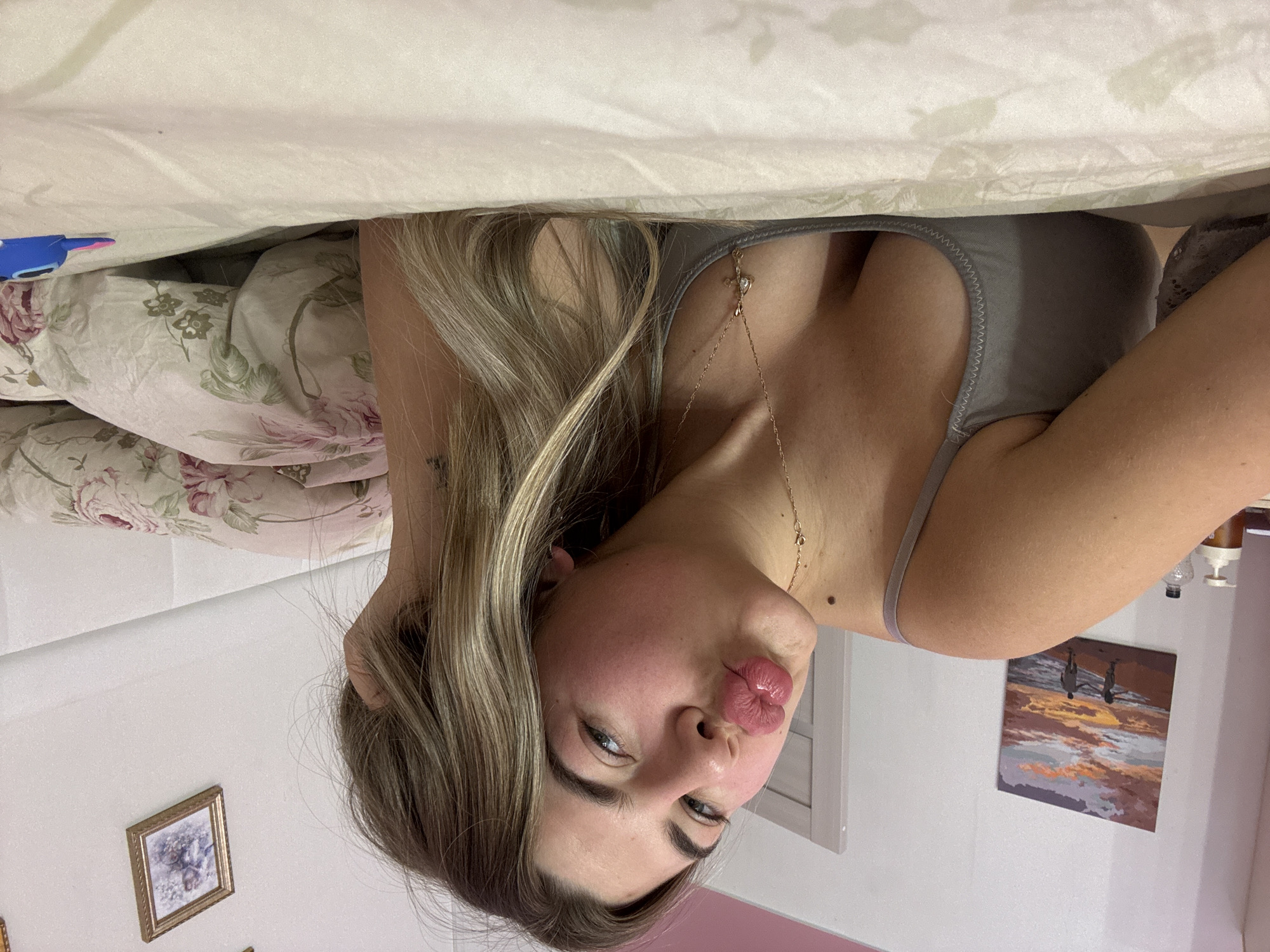 A woman is lying in bed. She has long, blonde hair and is wearing a gray tank top. She has her lips puckered. Her right hand is in her hair. There is a picture on the wall with two people standing, and one of a landscape. The bedding is floral print. The photograph was taken indoors in a room that seems to be in Chaikovsky, Russia.
