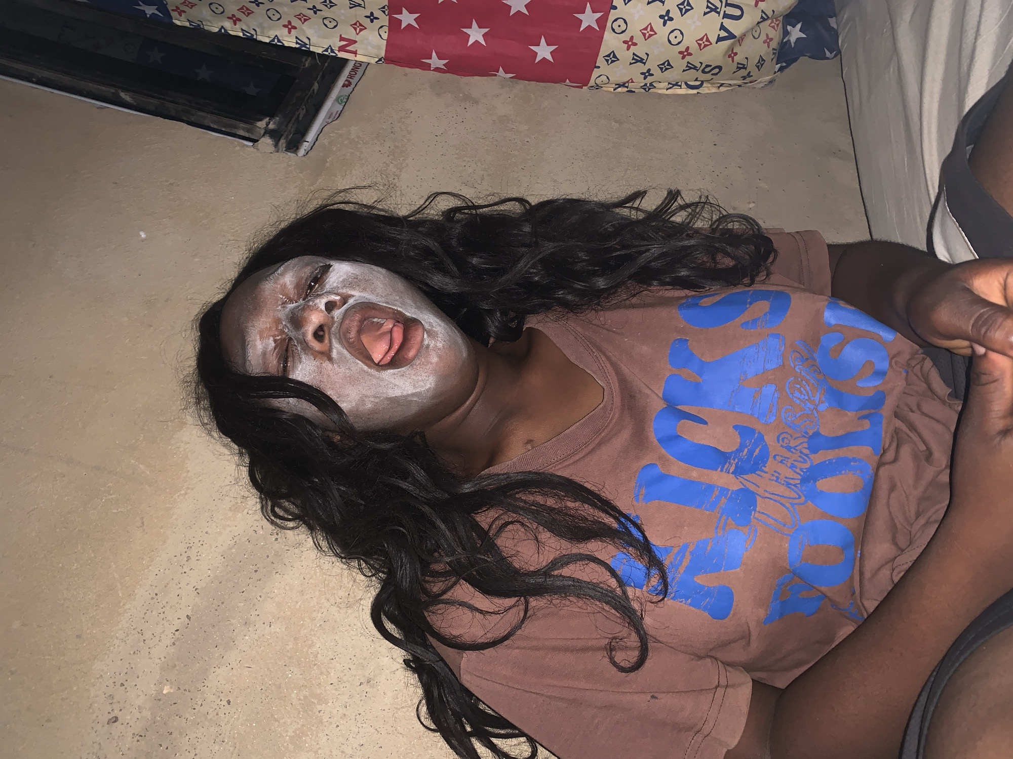 A Black woman lies supine on a pale tan surface, possibly a floor, with a white facial mask applied to her face. Her mouth is open, exposing her tongue. She is wearing a brown t-shirt with blue lettering. She has long, wavy black hair. In the background, there is a patterned pillow and a glimpse of a bed. A male arm and hand with gray and black clothes is in the lower right corner. The location is potentially inside a residence in the city of Agban, Nigeria.