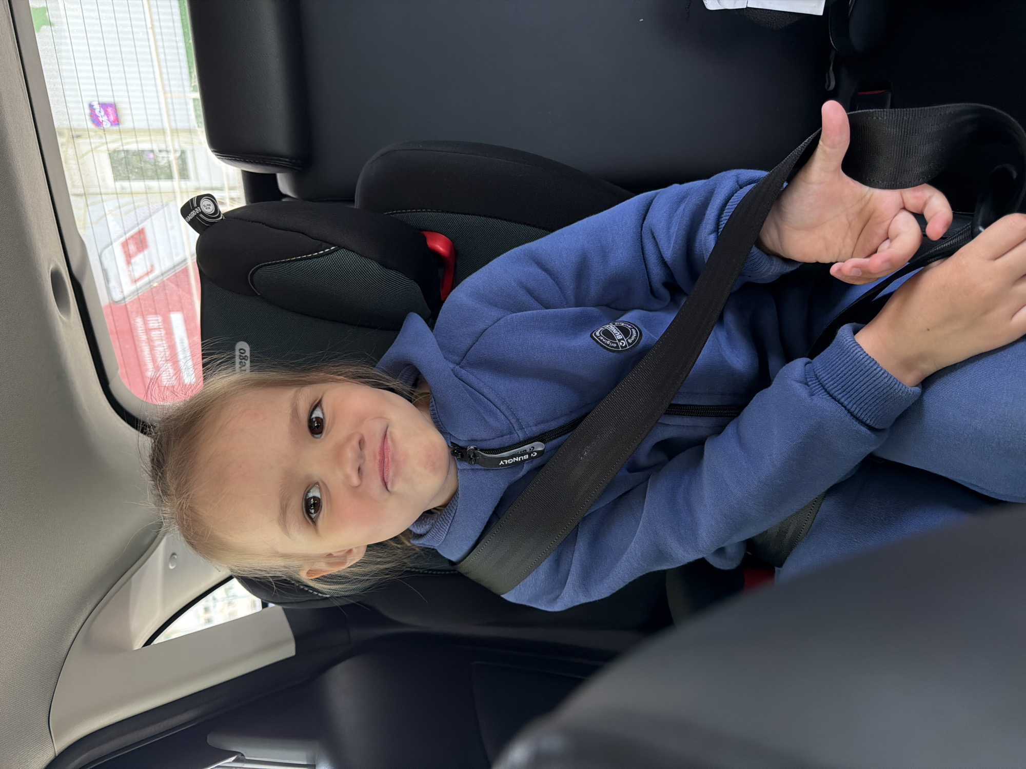 A young girl is seated in a car child seat, wearing a blue sweatshirt. She is strapped in with a seatbelt and giving a "thumbs up" gesture with her left hand. The image appears to be taken from within the car. Through the window, elements of the outside world can be seen, including a building with red and white coloring and a sign in Russian. The location is Chaikovsky, Russia.