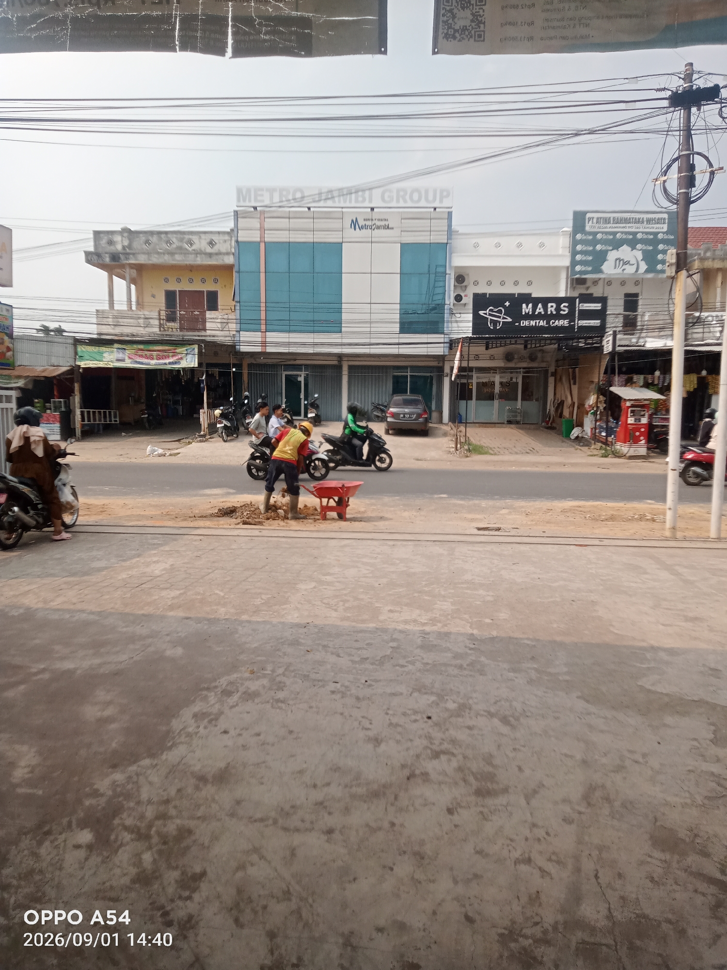 This is a street-level photograph taken in Jambi, Indonesia, showing a commercial area with several buildings. In the foreground, there is a gray concrete surface, possibly a sidewalk or street. Further back, a street runs horizontally across the image. On the street, a worker in a yellow vest and blue pants is using a red wheelbarrow and shovel to move dirt, suggesting construction or road repair. Several motorcycles are parked or in motion on the street, and a few people are visible, including a woman on a motorcycle in the left foreground and other individuals on motorcycles in the background.
The buildings in the background display various architectural styles and signage. One prominent building features a modern facade with large blue glass windows and a white section with the text "METRO JAMBI GROUP" and "Metro Jambi". To its right is another building with a white facade and signage for "PT. ATINA RAHMATAKA WISATA" and a smaller sign that says "MARS - DENTAL CARE -". Further to the left, there are older-looking buildings with awnings and signs, one of which appears to be a shop or market. Power lines are visible crisscrossing the sky above the buildings. The overall impression is of a busy urban environment.