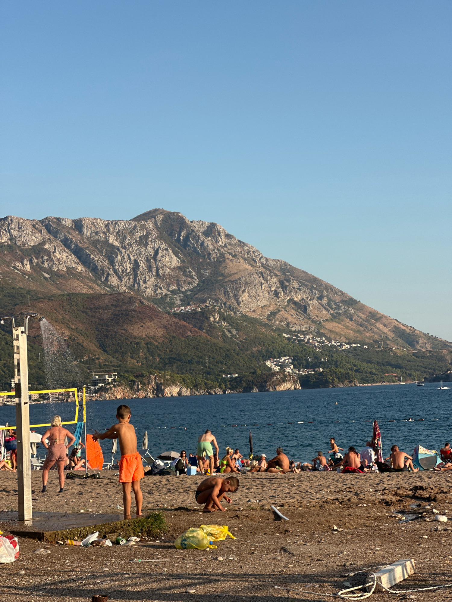 A beach scene in Boreti, Montenegro, features a group of people enjoying a sunny day by the Adriatic Sea, with a rugged mountain range serving as a dramatic backdrop. The water is a deep blue, and a clear sky suggests fair weather.
Several individuals are scattered across the sandy beach. In the foreground, a young boy in orange shorts stands near a shower unit, his arms outstretched. To his left, a woman in a pink outfit is also near the shower. Other beachgoers are visible lounging on chairs, sitting on the sand, and walking. Some are in swimsuits, suggesting a typical beach day. A volleyball net is set up on the sand.
The beach is somewhat littered with various pieces of trash, including plastic bags, bottles, and wrappers, detracting from the otherwise idyllic setting. The sun appears to be setting or is in the late afternoon, casting long shadows and creating a warm, golden light on the scene. The mountains in the background are dry and rocky, with scattered greenery and a small settlement visible on their slopes. The overall atmosphere is one of relaxed leisure, despite the visible litter. The word "COPAVA" is visible on the volleyball net.