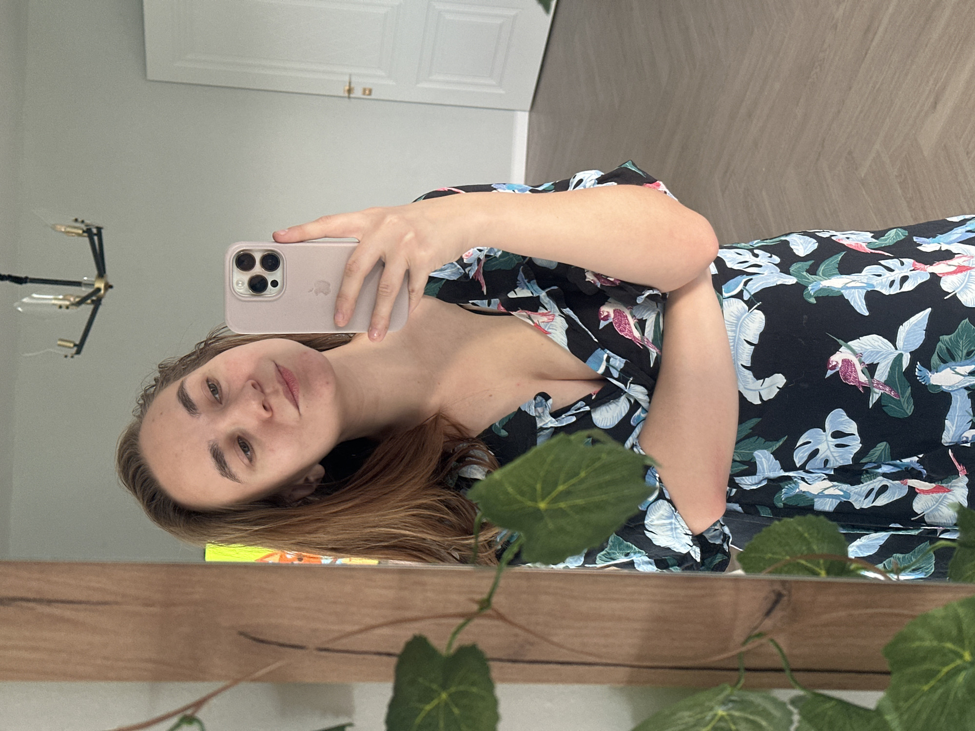 A woman with fair skin and brown hair is taking a selfie in a mirror. She is holding a pink iPhone in her right hand. She is wearing a black floral dress with blue, white, and green botanical patterns. The mirror is framed by a wooden structure, and a leafy plant is visible in the foreground. In the background, there is a door and a wall, with a modern chandelier visible on the left. There is also a herringbone patterned floor. There is no information to confirm this image was taken in Kharnavy, Russia.