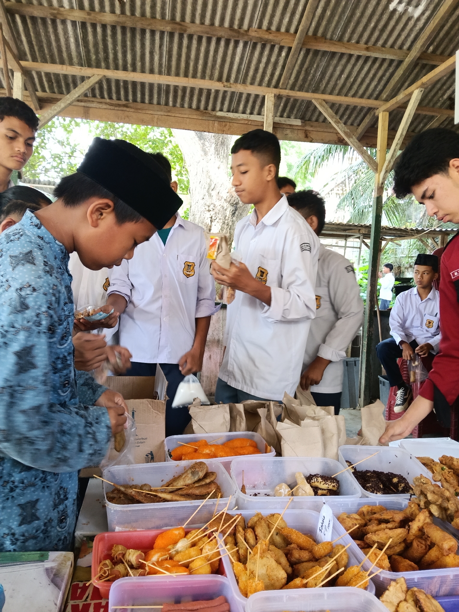 The image shows a group of boys in Lhokseumawe, Indonesia, gathered around a street food vendor. They are dressed in school uniforms, mostly white shirts with a patch on the left side, and black pants. One boy is wearing a blue patterned shirt and a black cap. They are interacting with the vendor, who is out of frame. In front of them, several plastic containers display various fried snacks on sticks. These snacks include items wrapped in batter, fried fish, and sausages. The vendor is packaging some of these items in brown paper bags. The scene is under a corrugated metal roof supported by wooden beams. In the background, there is a glimpse of greenery and another boy sitting on a stool.