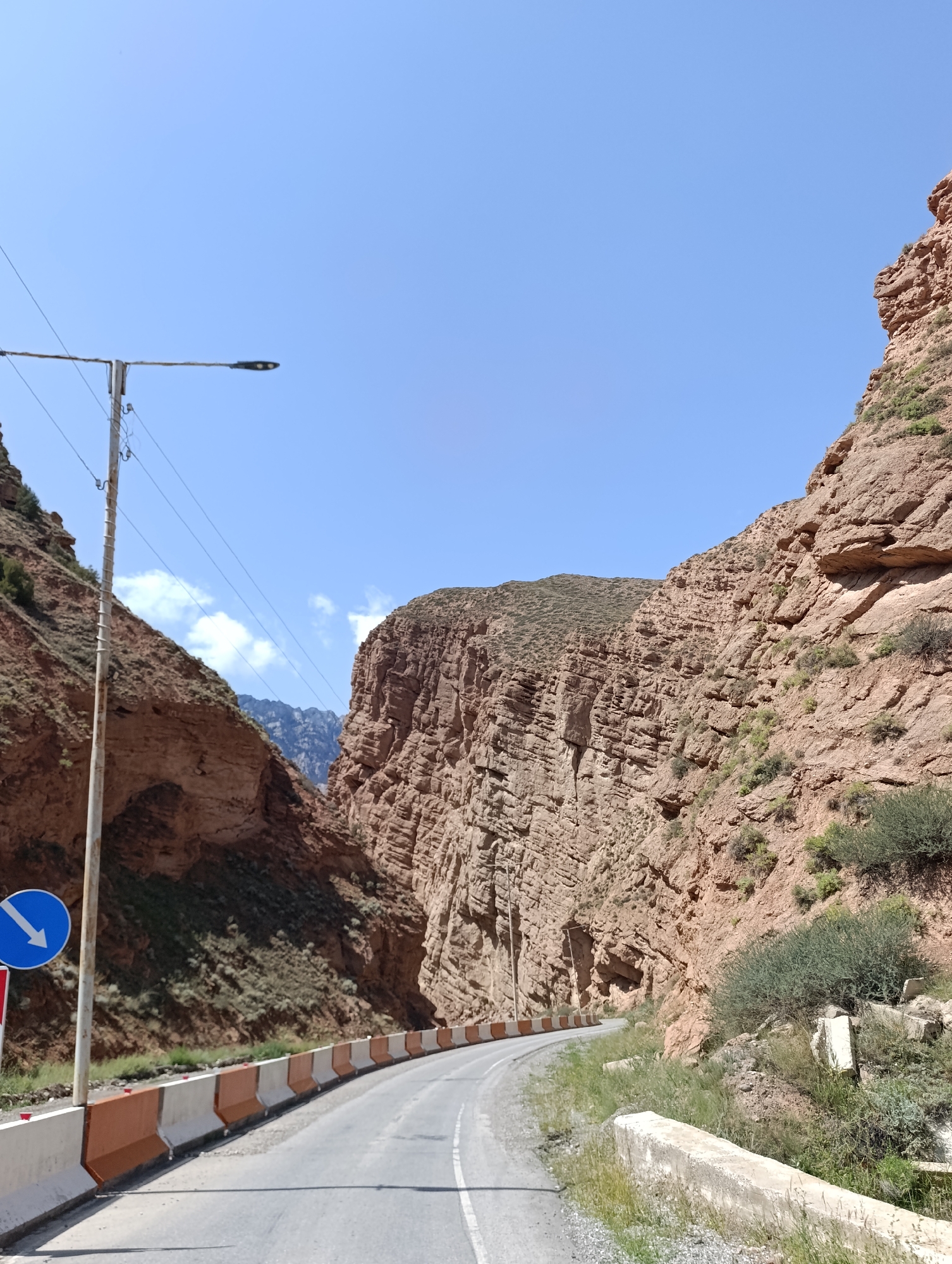The image captures a scenic view of a winding road passing through a dramatic mountain gorge. The road is paved and has lane markings. On the left side of the road, a street light stands tall, with a blue circular sign depicting a white arrow pointing downwards to the left. Orange and white concrete barriers line the edge of the road, separating it from the rocky terrain. The gorge walls are steep and made of layered, reddish-brown rock, with sparse vegetation growing on their slopes. In the distance, a mountain range can be seen under a clear blue sky with a few scattered white clouds. The overall scene depicts a remote, natural landscape with a road built for passage through challenging topography.